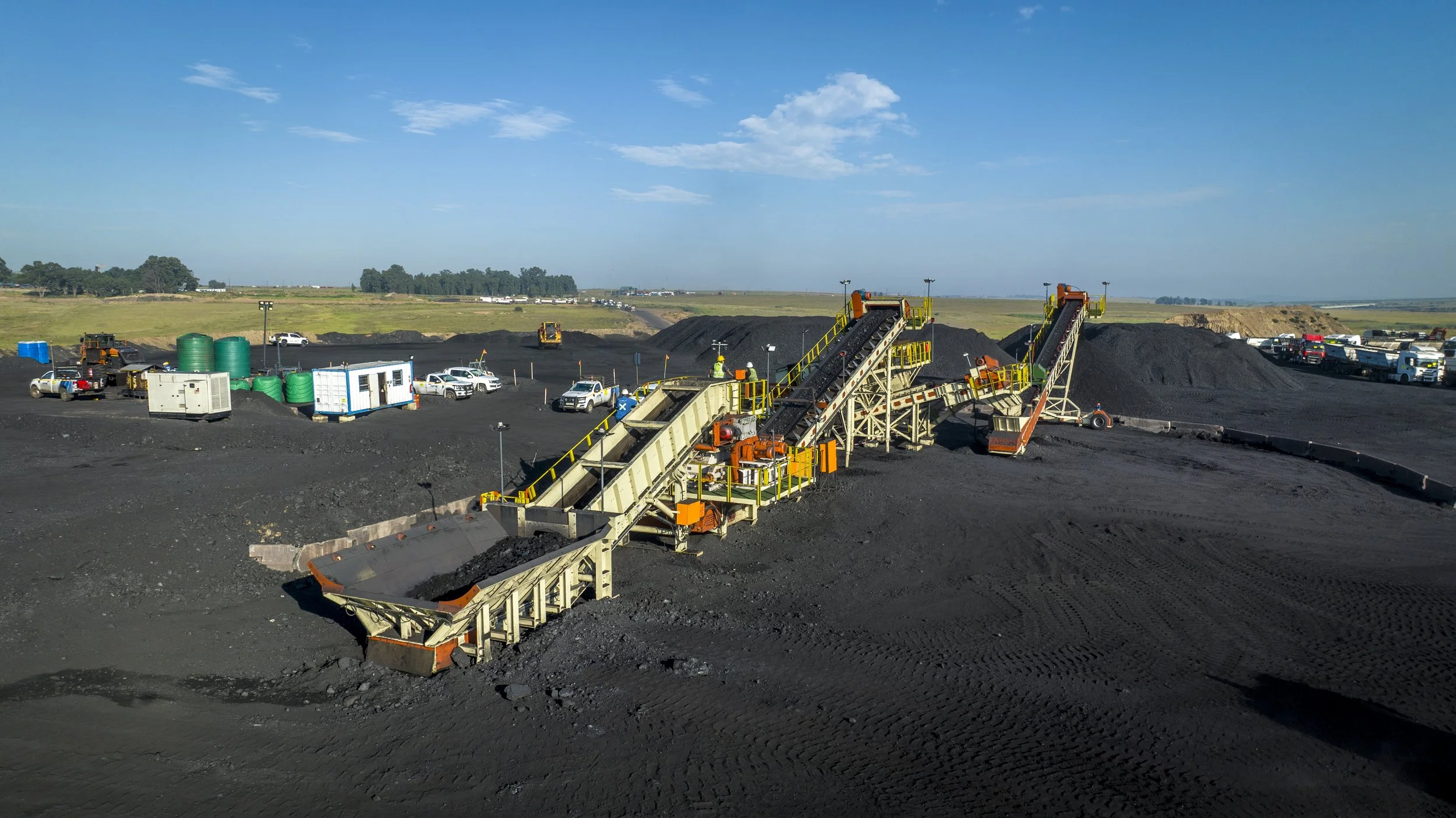 Open-pit mining operation with large conveyor belts transporting dark mineral material across black ground, surrounded by construction vehicles and equipment under a clear blue sky.