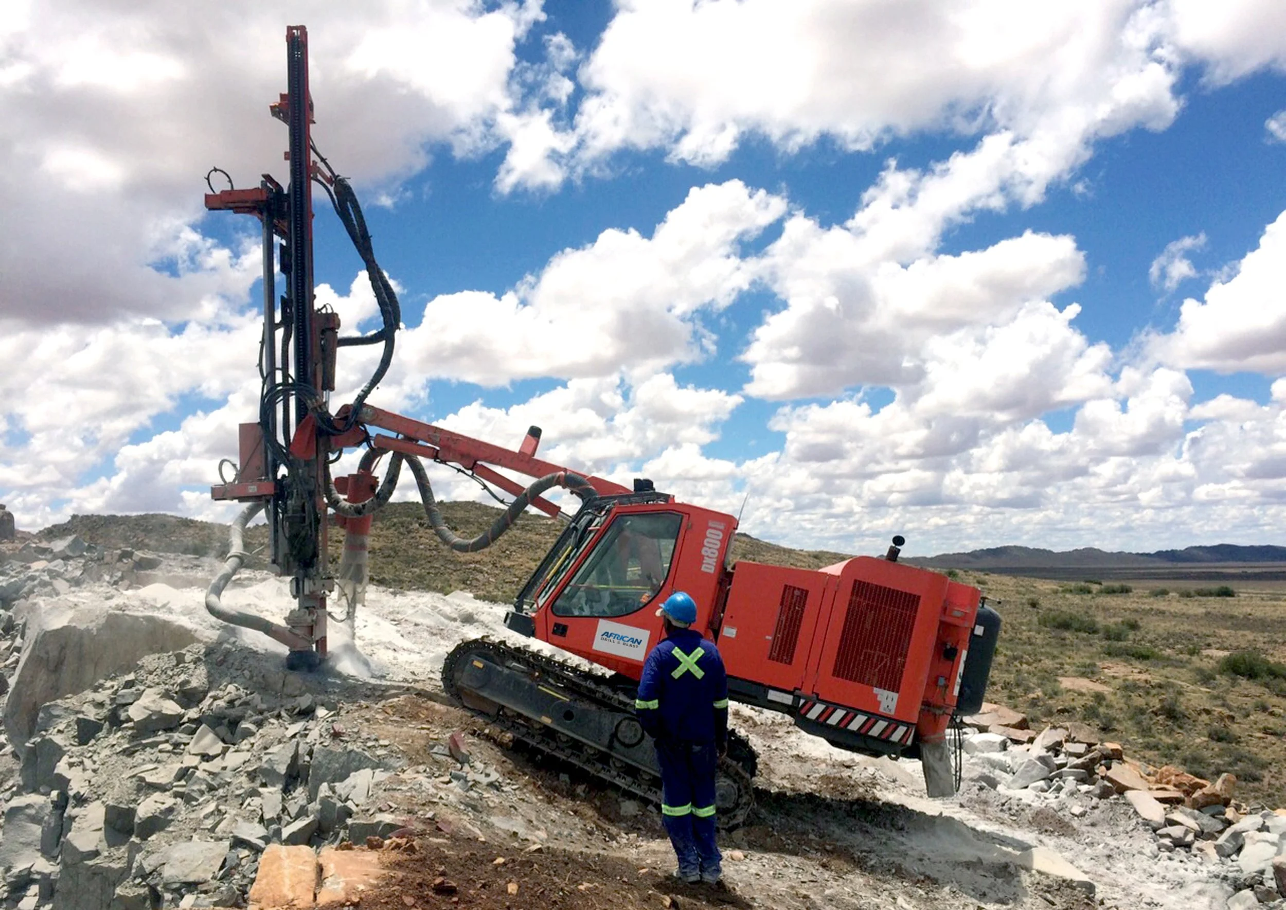 A worker in a blue safety uniform and helmet observes a large red industrial drilling machine mounted on tracks, working on rocky terrain under a partly cloudy blue sky.