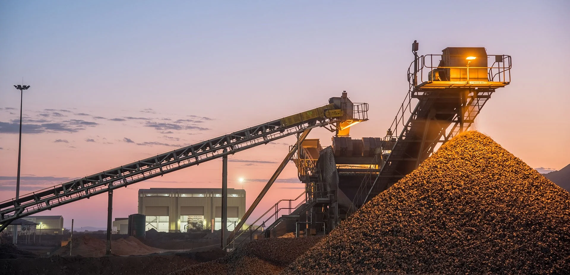 Mining operation at dusk with a conveyor belt transporting gravel into a large pile, and industrial buildings in the background.