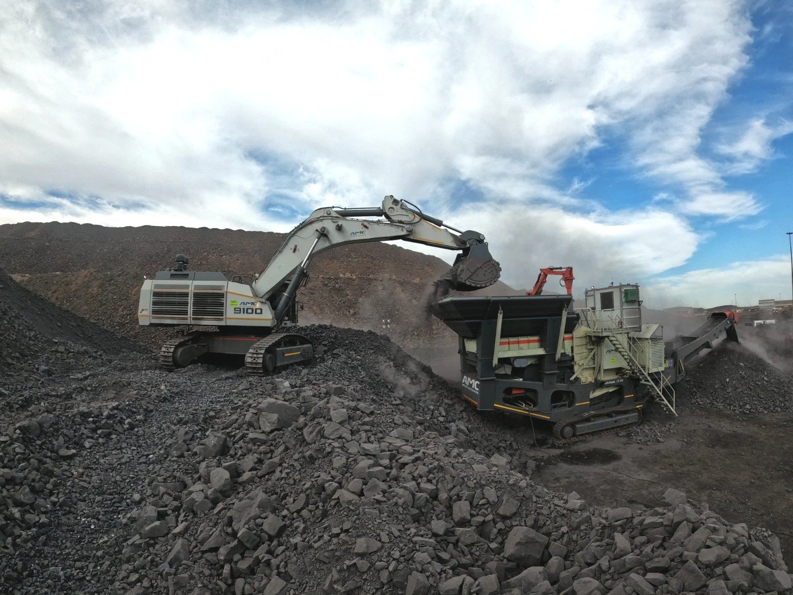 A large excavator with tracked wheels and a long arm is loading rocks and gravel into a mobile crusher machine at a quarry, with a blue sky and white clouds overhead.