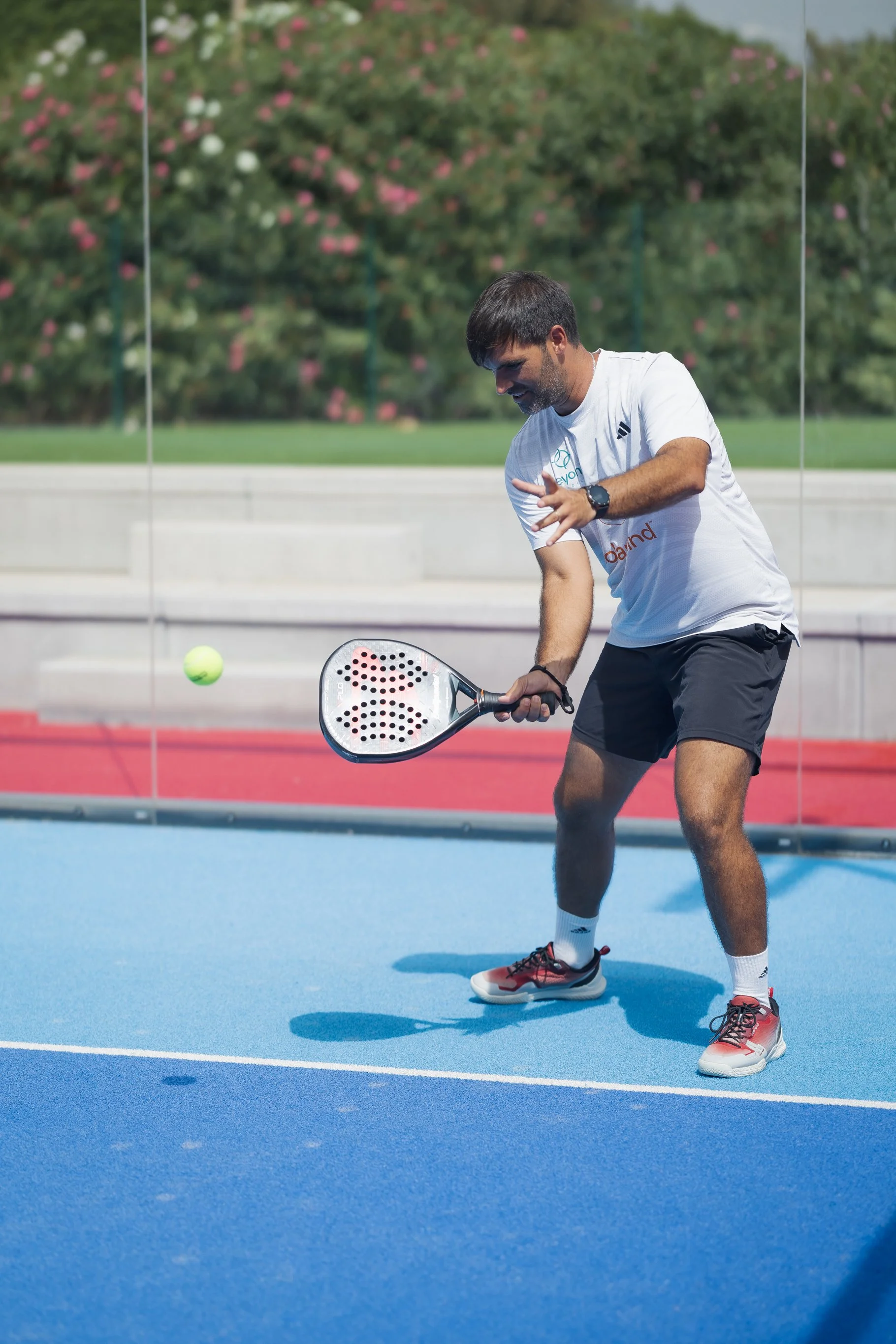 A man playing tennis on an outdoor court with a tennis ball in mid-air. He is dressed in a white shirt, black shorts, and red and white Nike shoes, holding a tennis racket, and appears to be preparing to hit the ball.