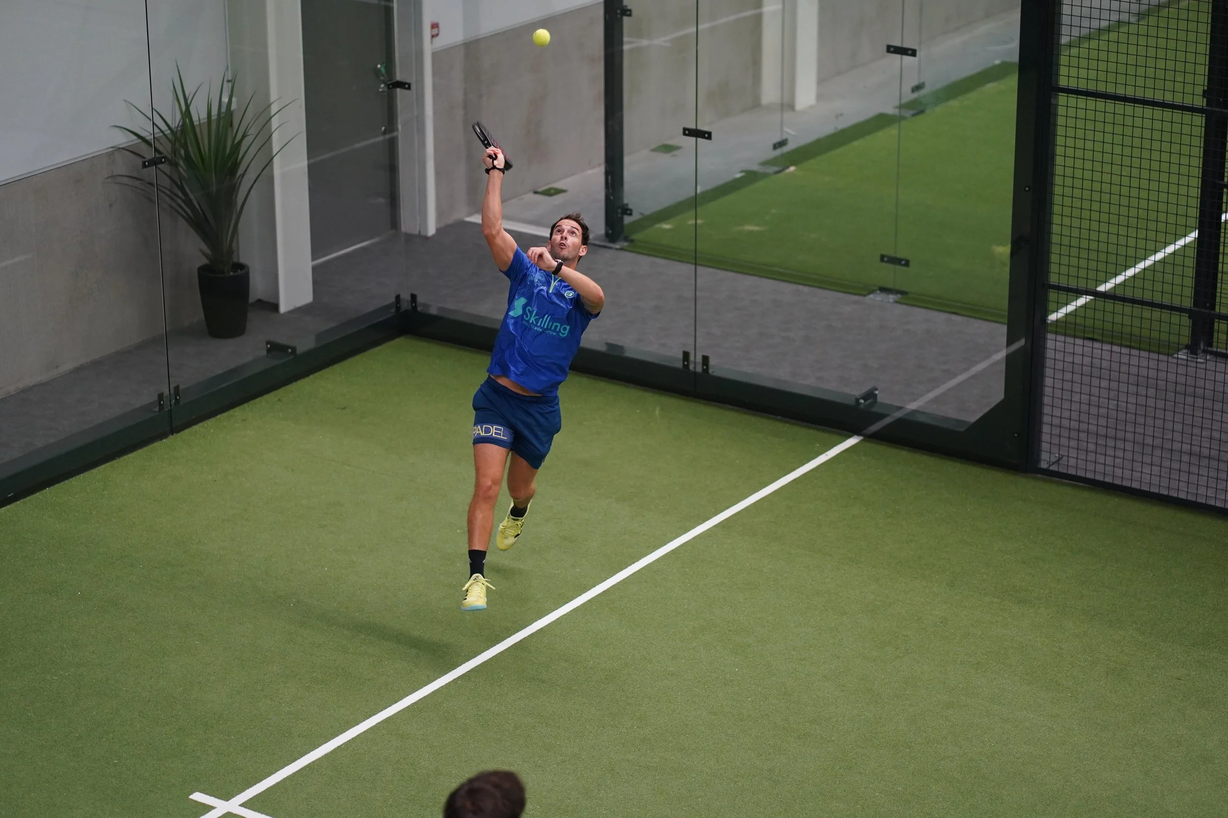A man playing paddle tennis on an indoor court, dressed in blue sports attire, hitting a yellow ball with a paddle.