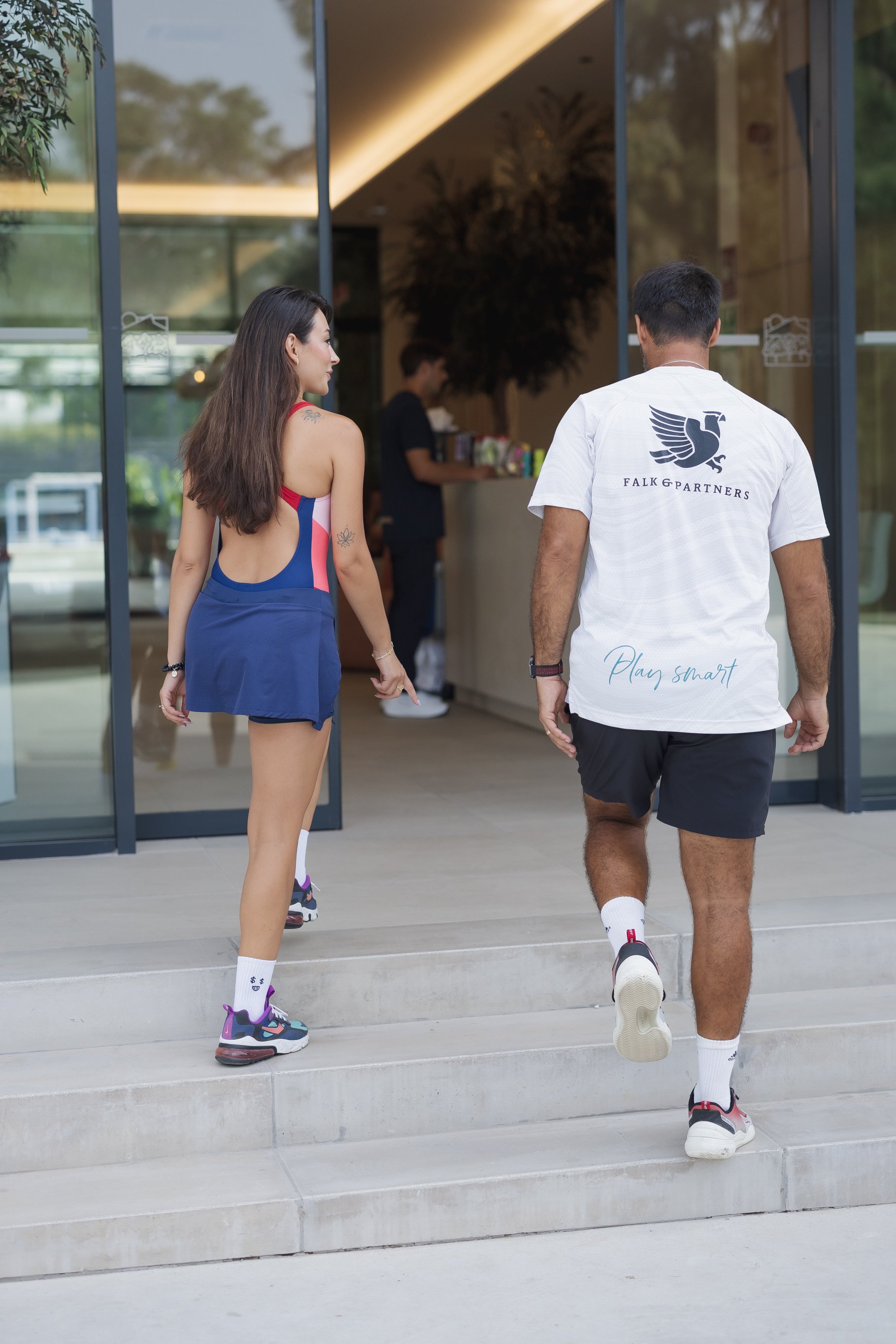 A man and a woman are walking up the stairs outside a modern building with large glass windows. The woman has long brown hair and tattoos, wearing a sports outfit, and the man has short dark hair, wearing a white t-shirt with a black eagle logo and b