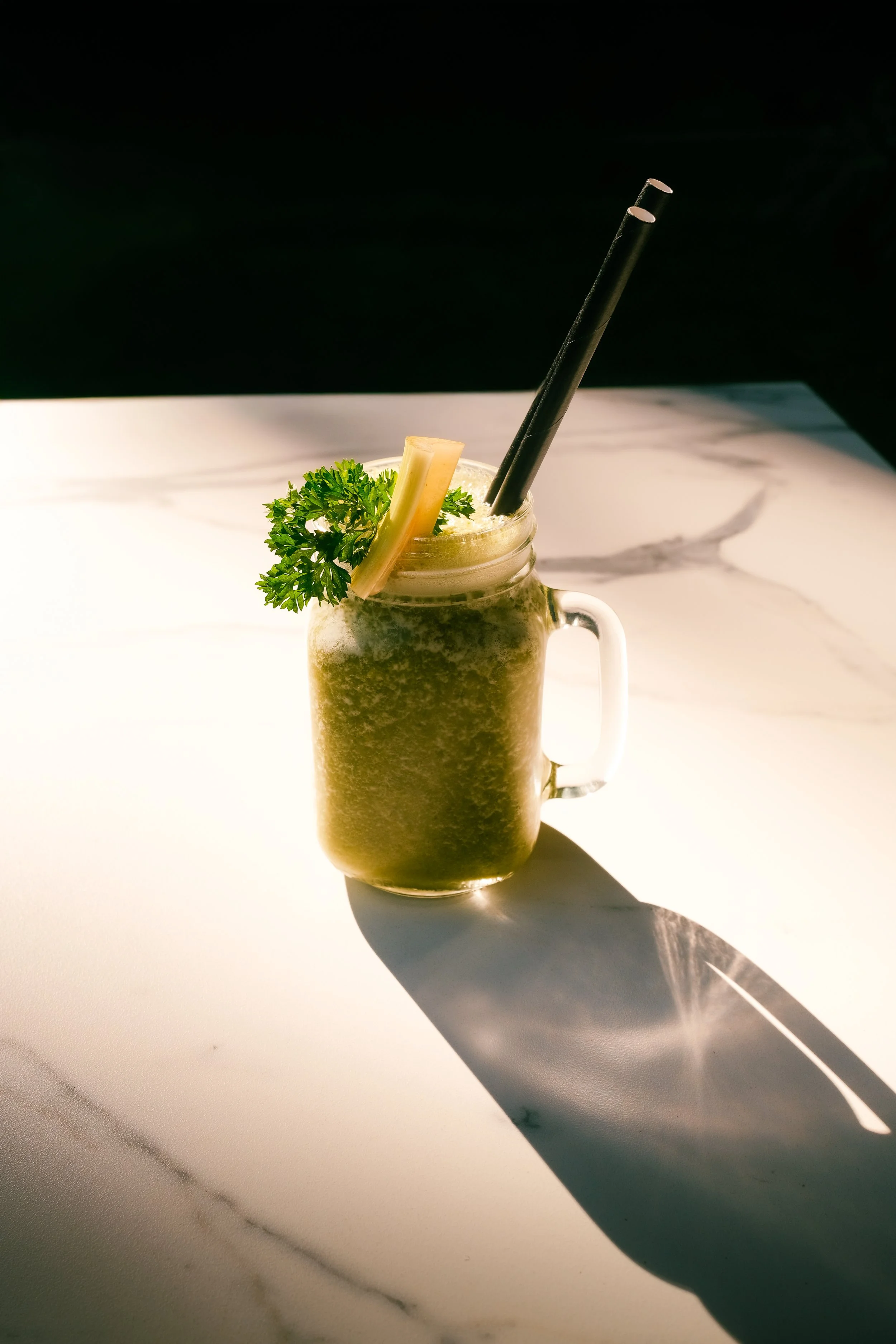 A jar filled with a green smoothie, garnished with a sprig of parsley and a pineapple chunk, with two black straws inside, placed on a white marble surface with a dark background.