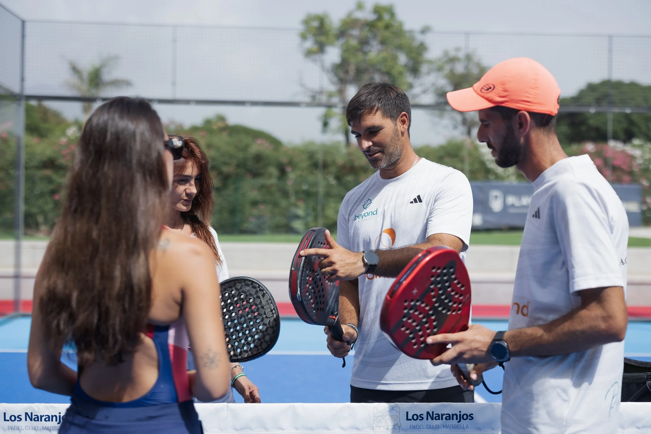 Group of people talking on a tennis court, some holding padel rackets, with a fence, trees, and blue sky in the background.