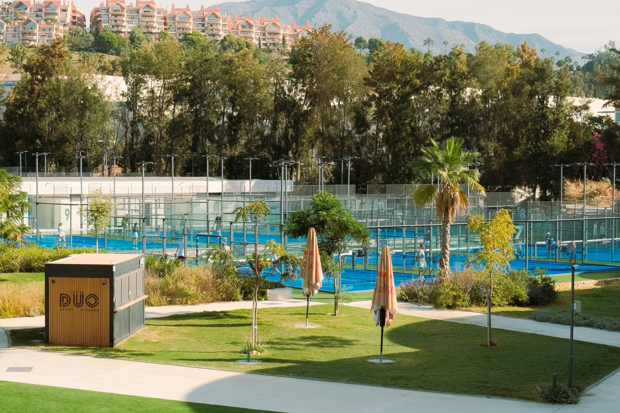 A tennis court with multiple players practicing, surrounded by greenery and trees, with residential buildings and hills in the background.