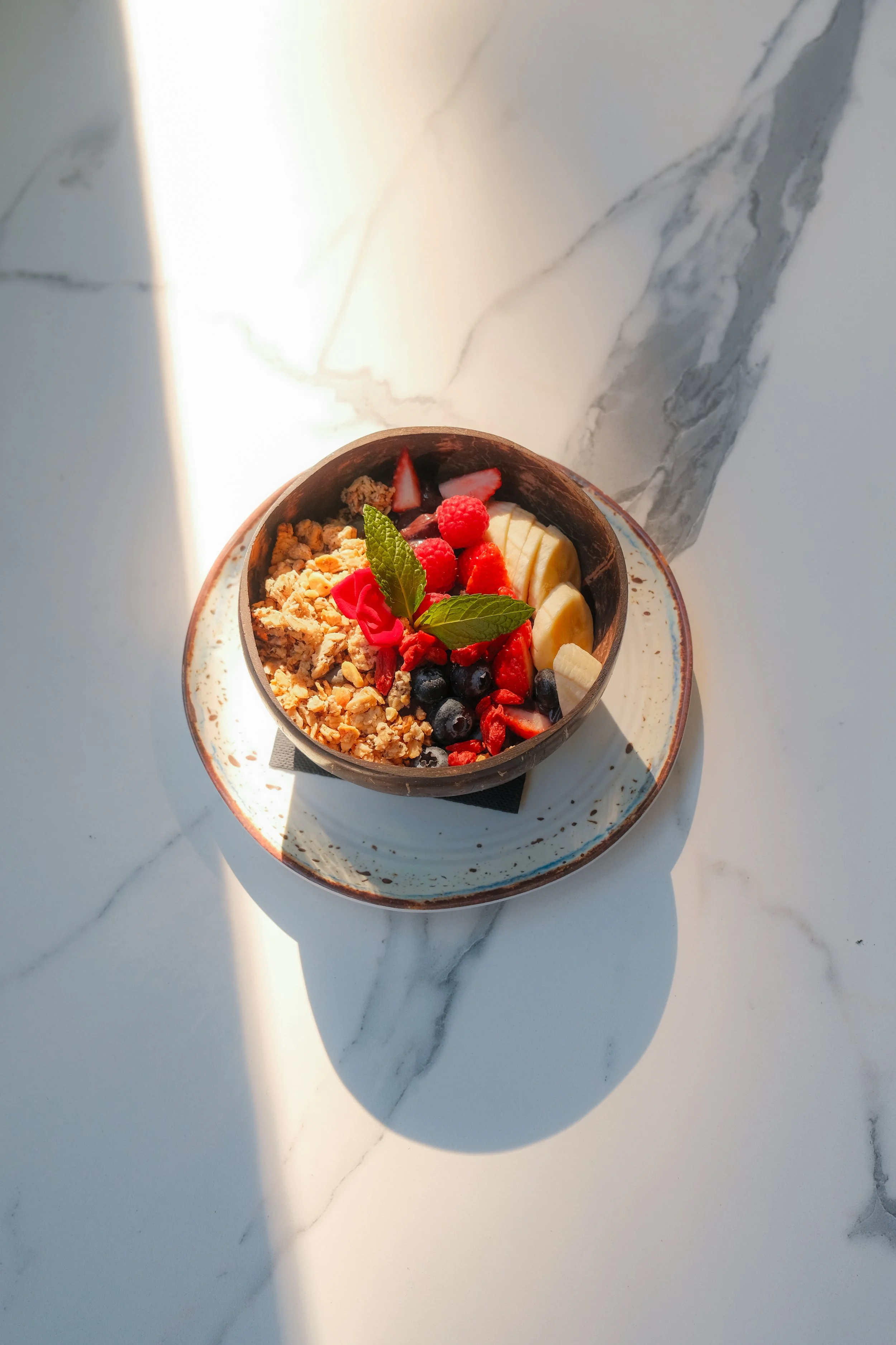A bowl filled with granola, sliced strawberries, blueberries, banana slices, and garnished with fresh mint leaves, placed on a white marble table with shadows and light.