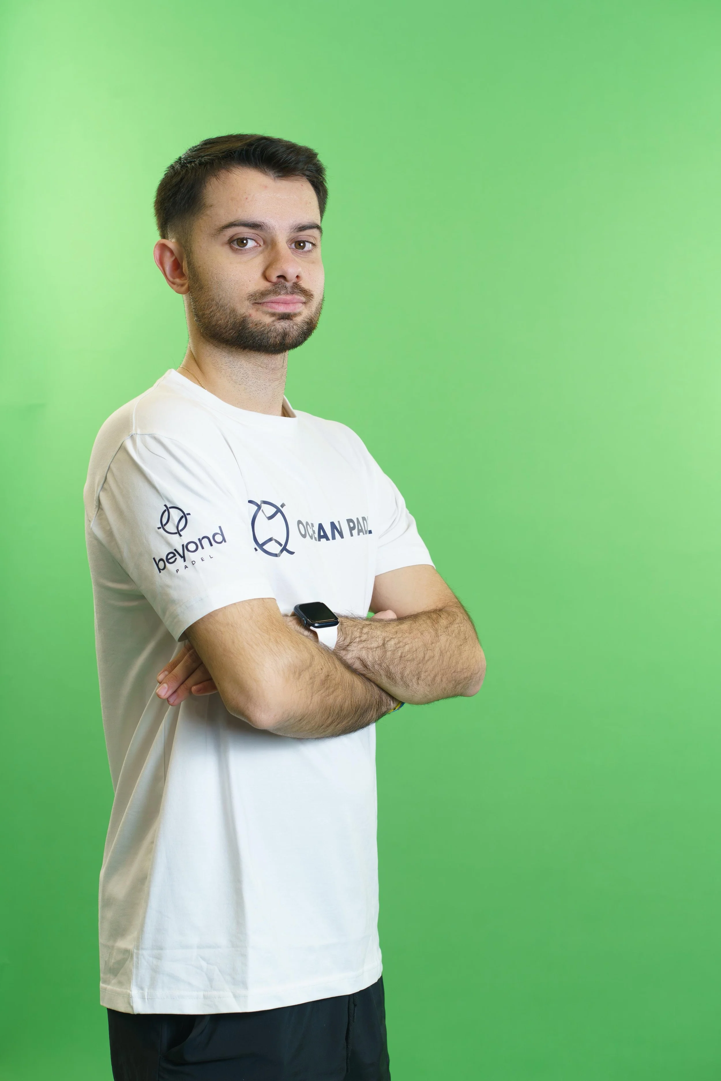 A young man with short dark hair and a beard standing with arms crossed in front of a green background. He is wearing a white t-shirt with logos and text that reads 'beyond OCEAN PADDLE' and a black watch on his left wrist.