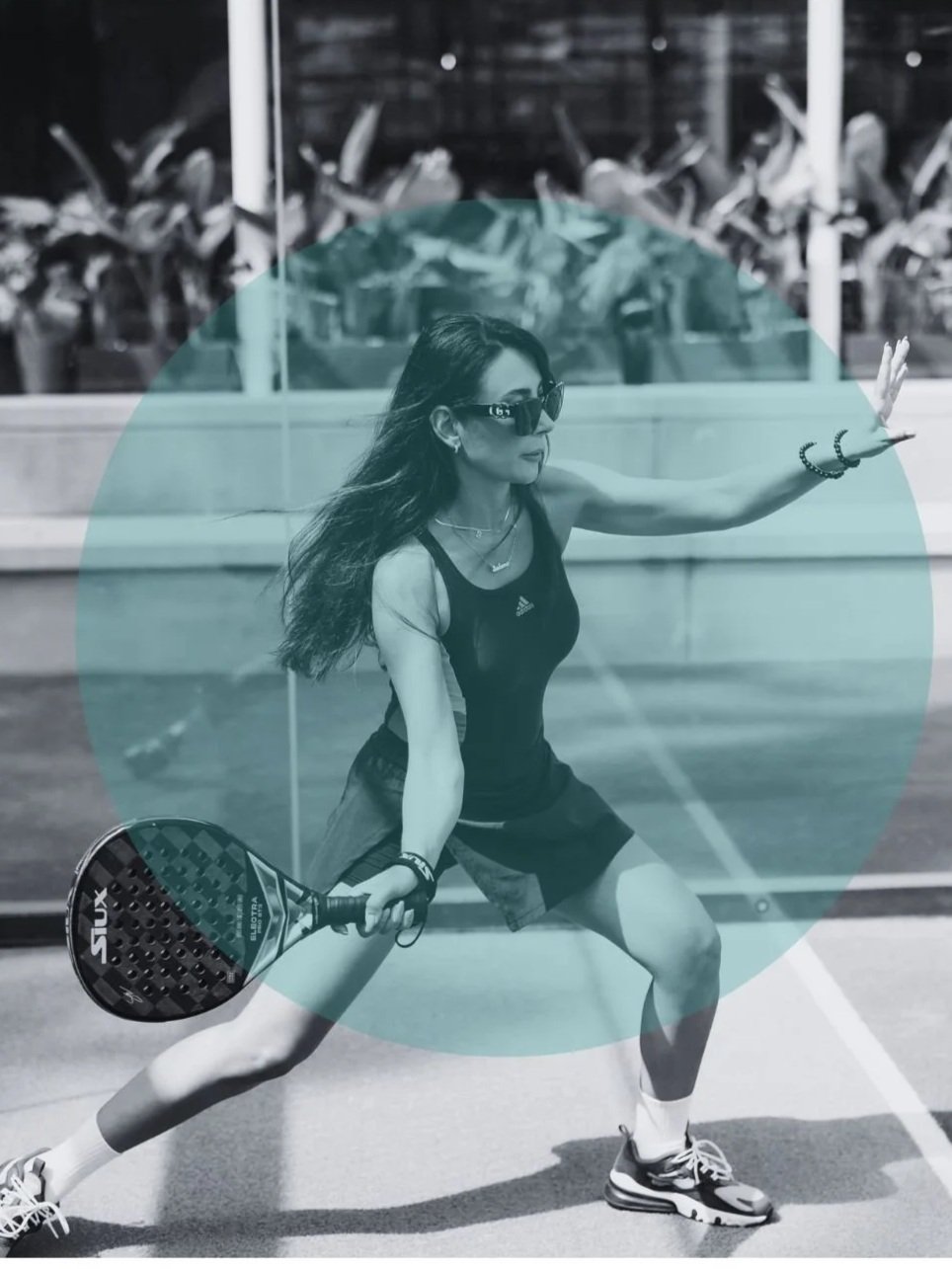 A woman playing pickleball outdoors, wearing sunglasses, a black athletic tank top, shorts, sneakers, and holding a pickleball paddle.