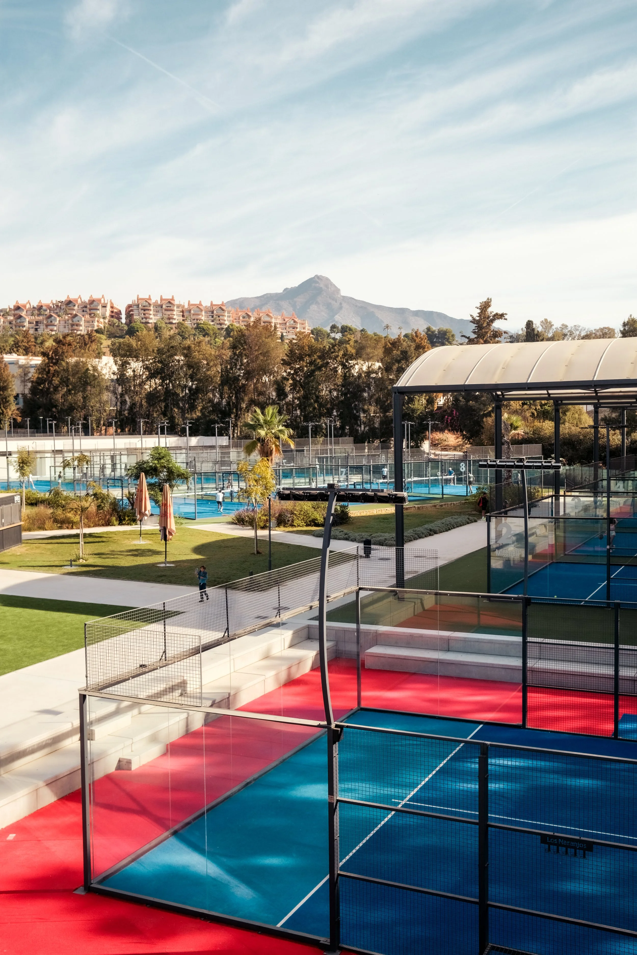 Outdoor sports complex with tennis courts, a covered basketball court, trees, a swimming pool, and residential buildings in the background with a mountain and blue sky.