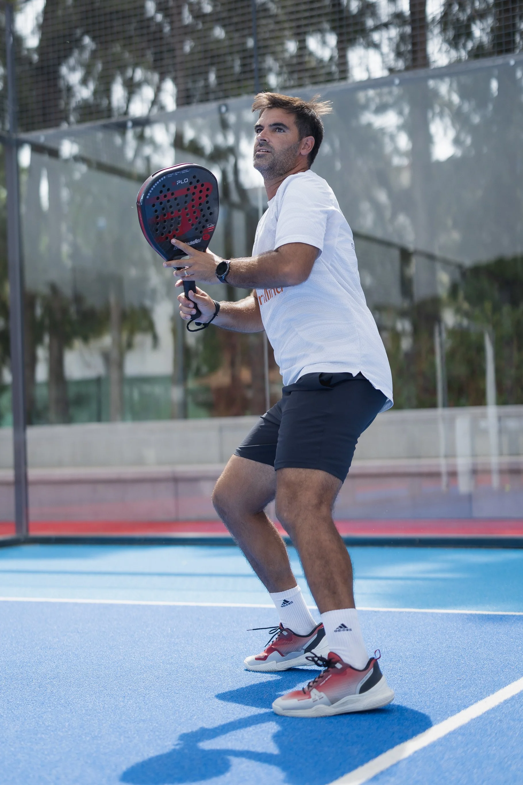 A man playing pickleball on a blue court with a black and red paddle, wearing white athletic shirt, black shorts, white socks, and red and white shoes, in an outdoor sports facility.