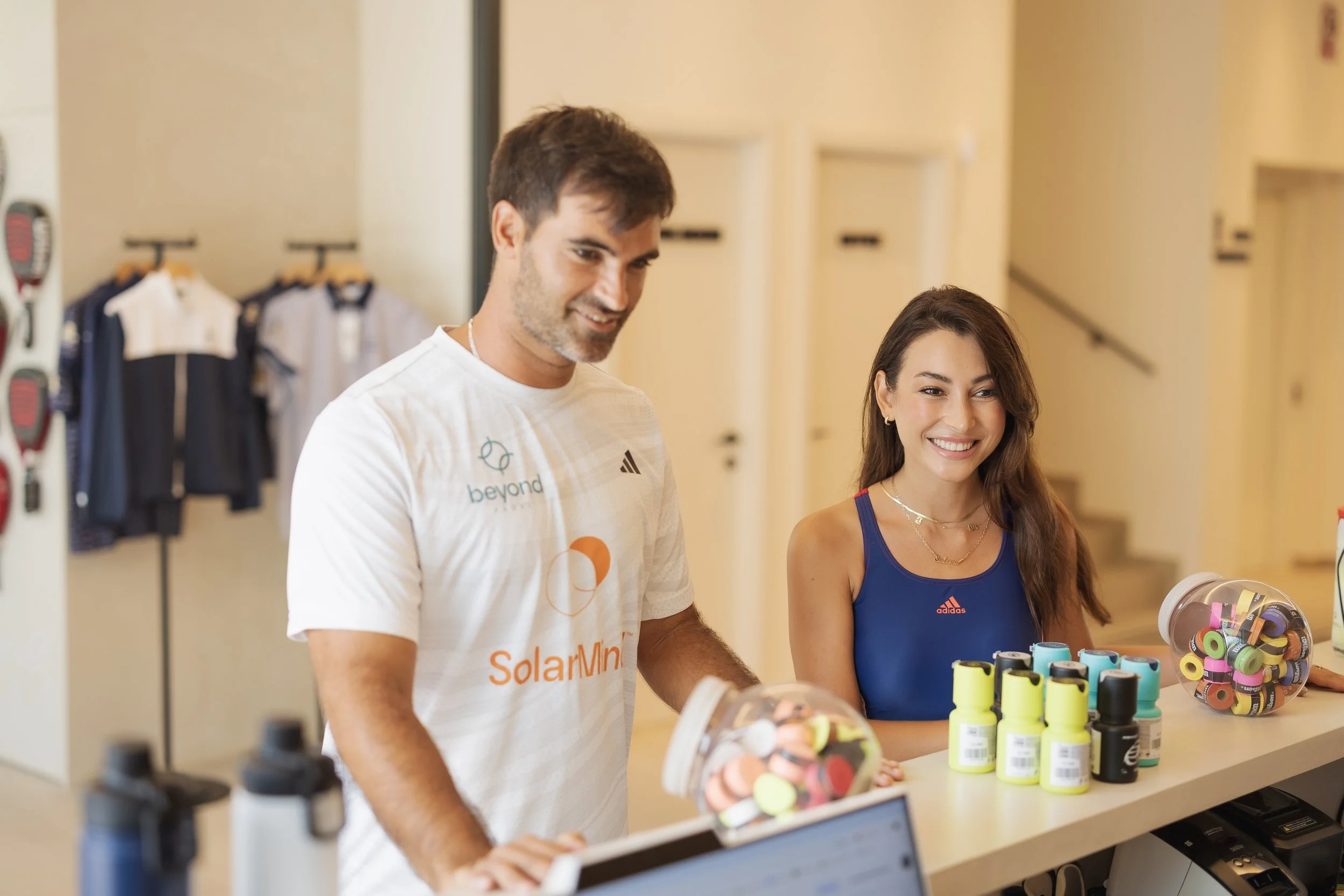 A man and woman are shopping for colorful wristbands at a store. The man is wearing a white T-shirt with logos, and the woman is in a blue sports top. They are smiling and looking at the wristbands displayed on the counter.
