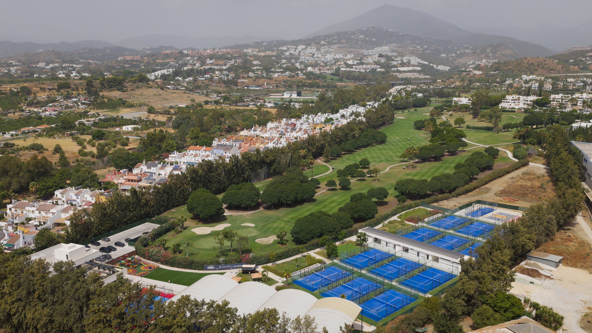 Aerial view of a cityscape featuring a golf course with multiple sand traps, tennis courts with blue surfaces, residential houses, a playground, and distant hills.