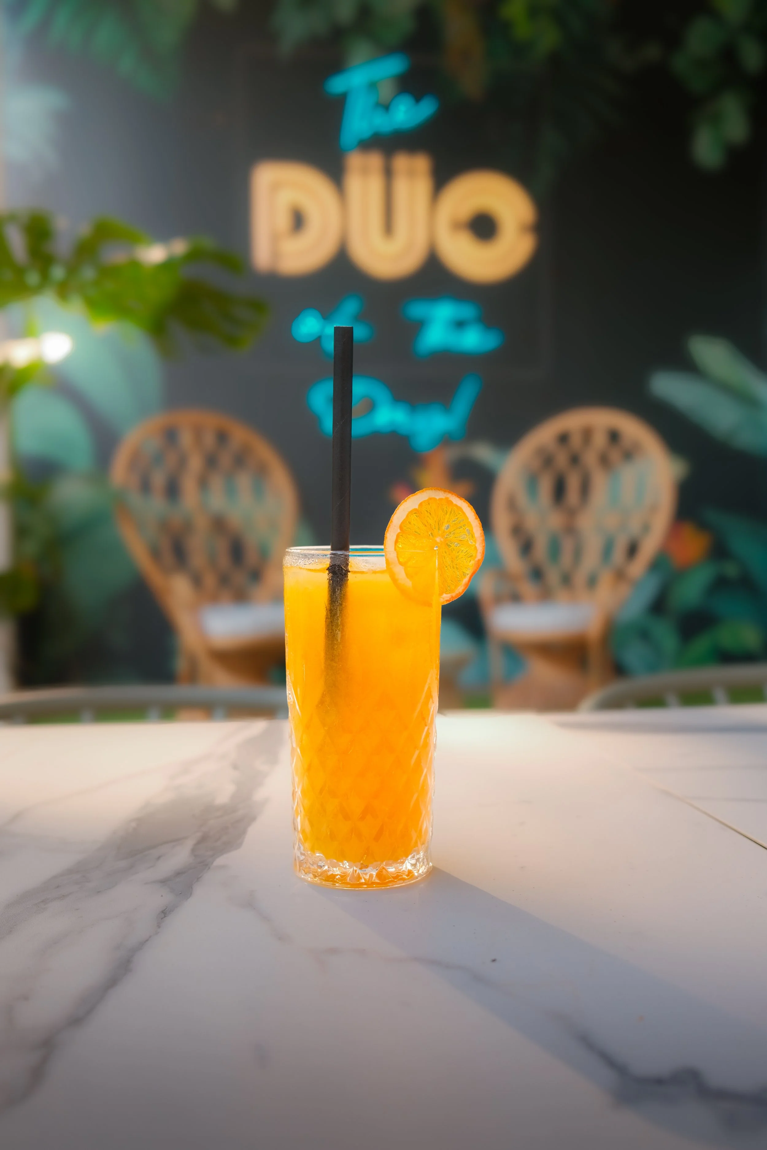 A tall glass of orange juice garnished with a slice of orange and a black straw, placed on a white marble table.