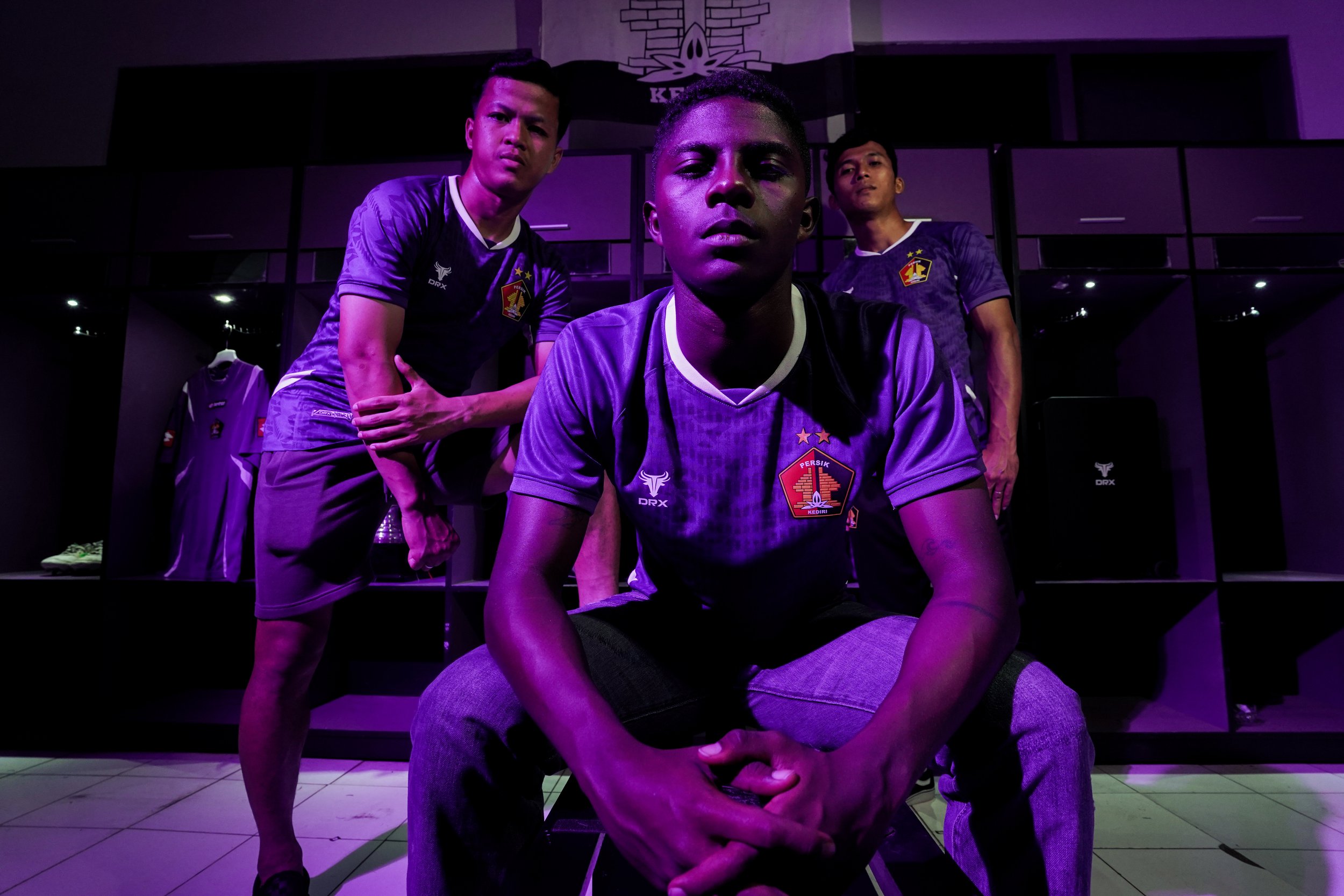 Three young men in purple soccer jerseys with a club crest pose in a locker room with purple lighting, black shelves and a soccer jersey on display.