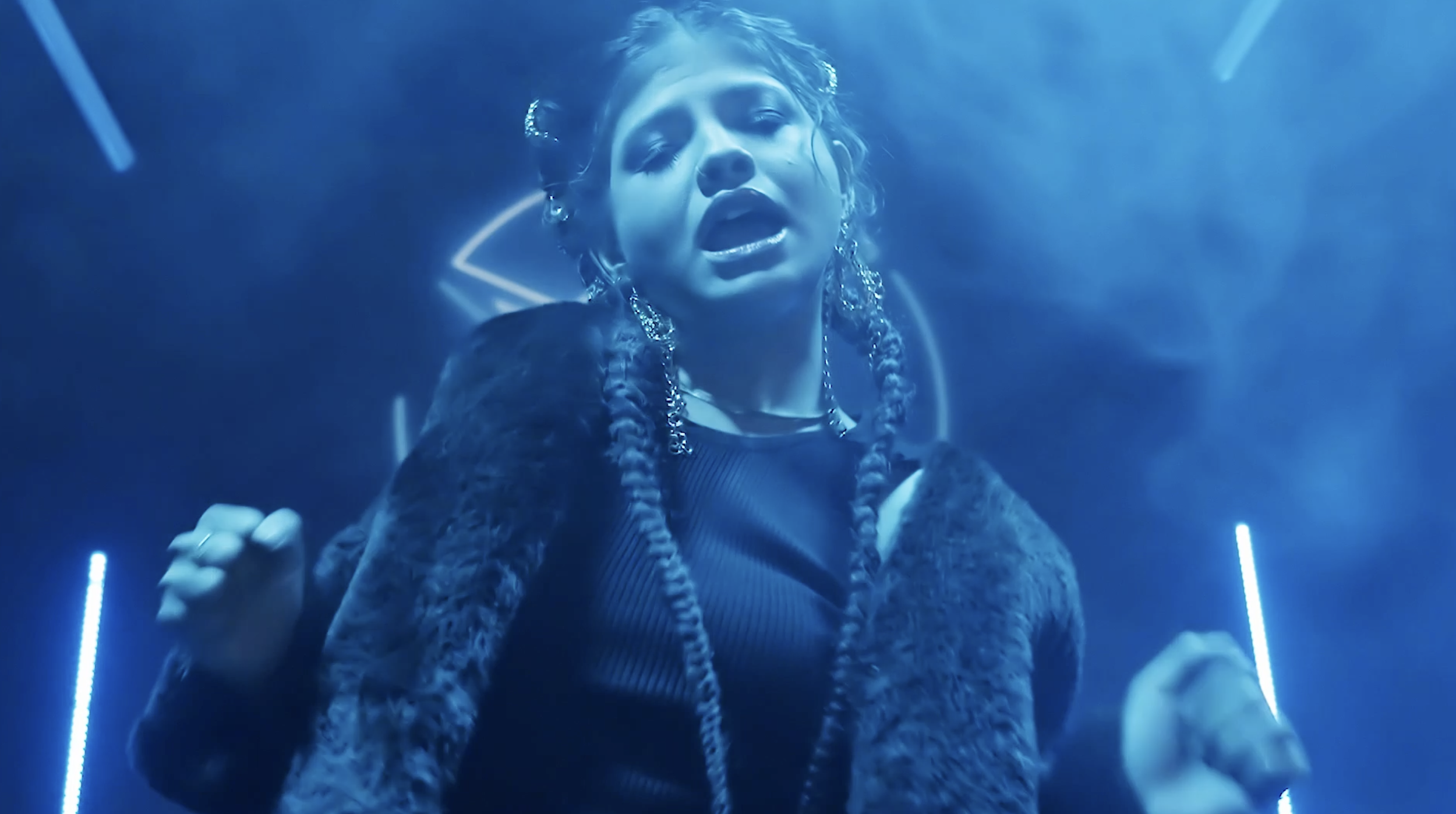 A woman with braids, wearing large earrings and a fur coat, appears to be singing or speaking passionately in a blue-lit setting.