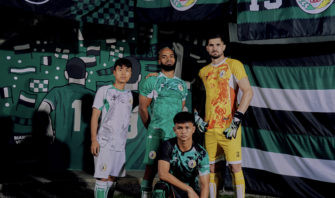 Four soccer players wearing jerseys with the Mexican flag colors, standing and kneeling in front of a mural with soccer themes and large Mexican flags.