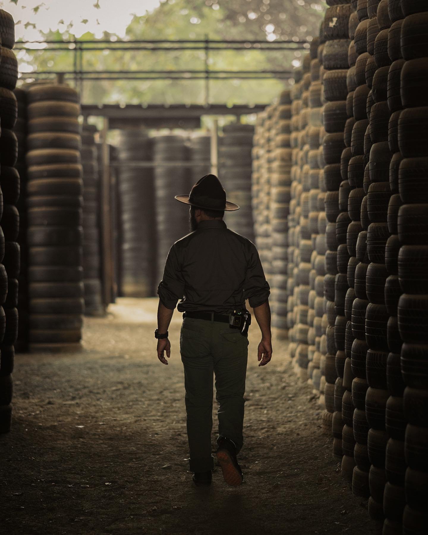 A person walking through a passageway with tall stacks of tires on either side, wearing a black shirt, green pants, and a wide-brimmed hat.