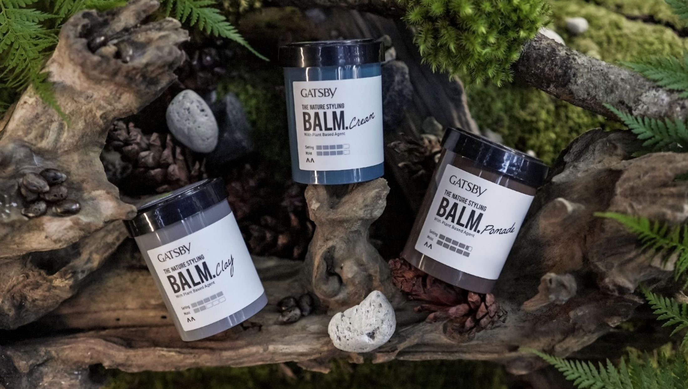 Three containers of Gatsby brand styling balms, labeled Clay, Cream, and Pomade, are placed on a natural wooden surface with rocks and greenery around them. The balms are in black jars with white labels.