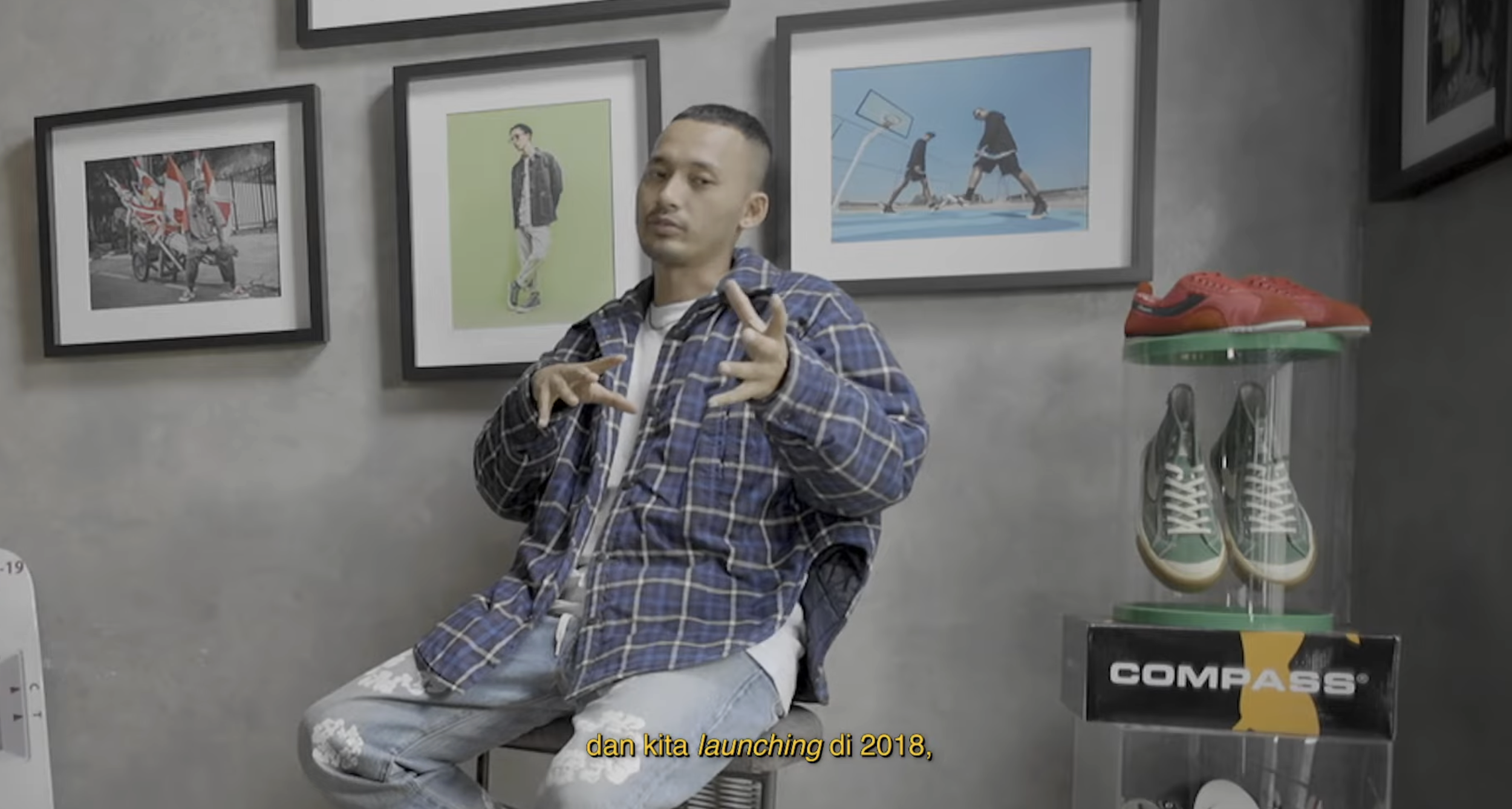 A man in a blue plaid jacket and ripped jeans sitting on a chair in a room with gray walls, surrounded by framed photos of skateboarders and sneaker displays.