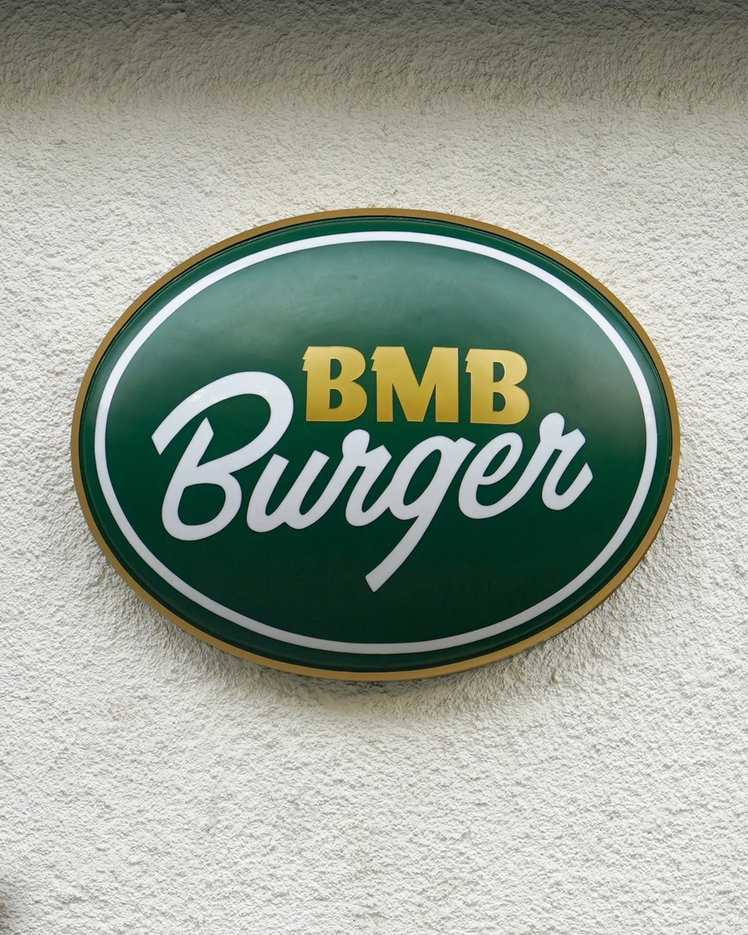 A green oval sign with gold and white text reading 'BMB Burger' mounted on a textured beige wall.
