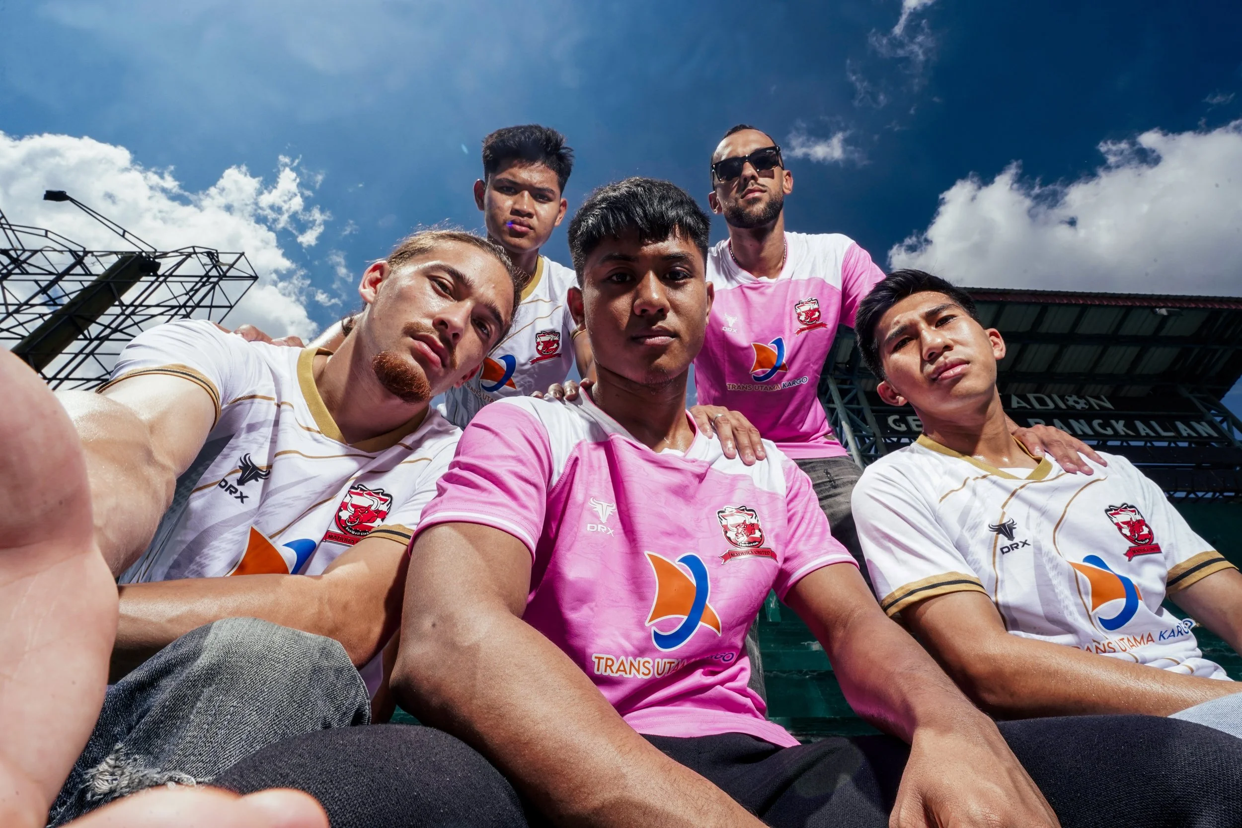 Group of six young men, some wearing soccer jerseys, on a stadium field, looking at the camera, under a blue sky with clouds.