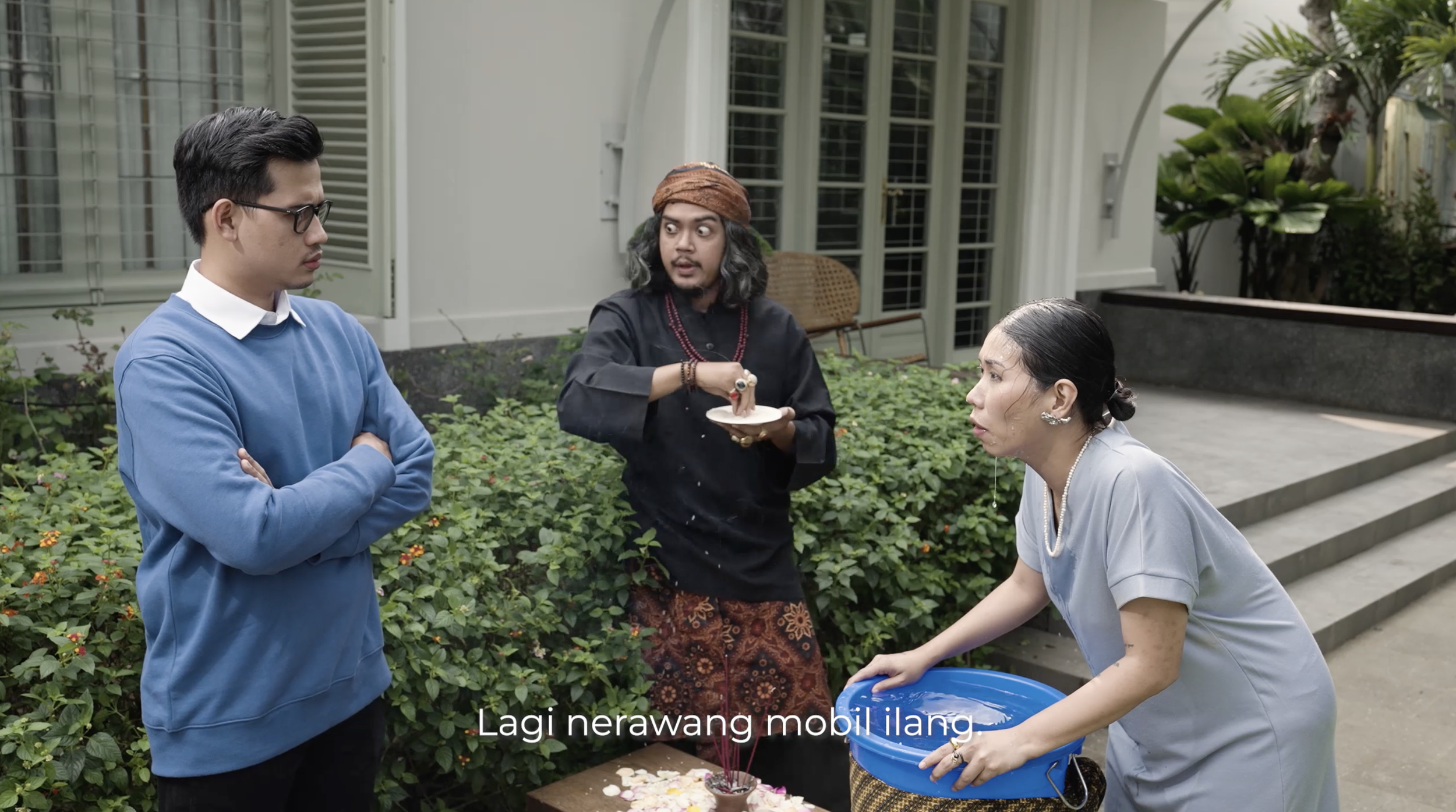 Three people engaged in a conversation outside a house with green plants. A woman is holding a blue bucket, a man with glasses and a blue sweater stands with arms crossed, and another person in black attire appears to be explaining something.
