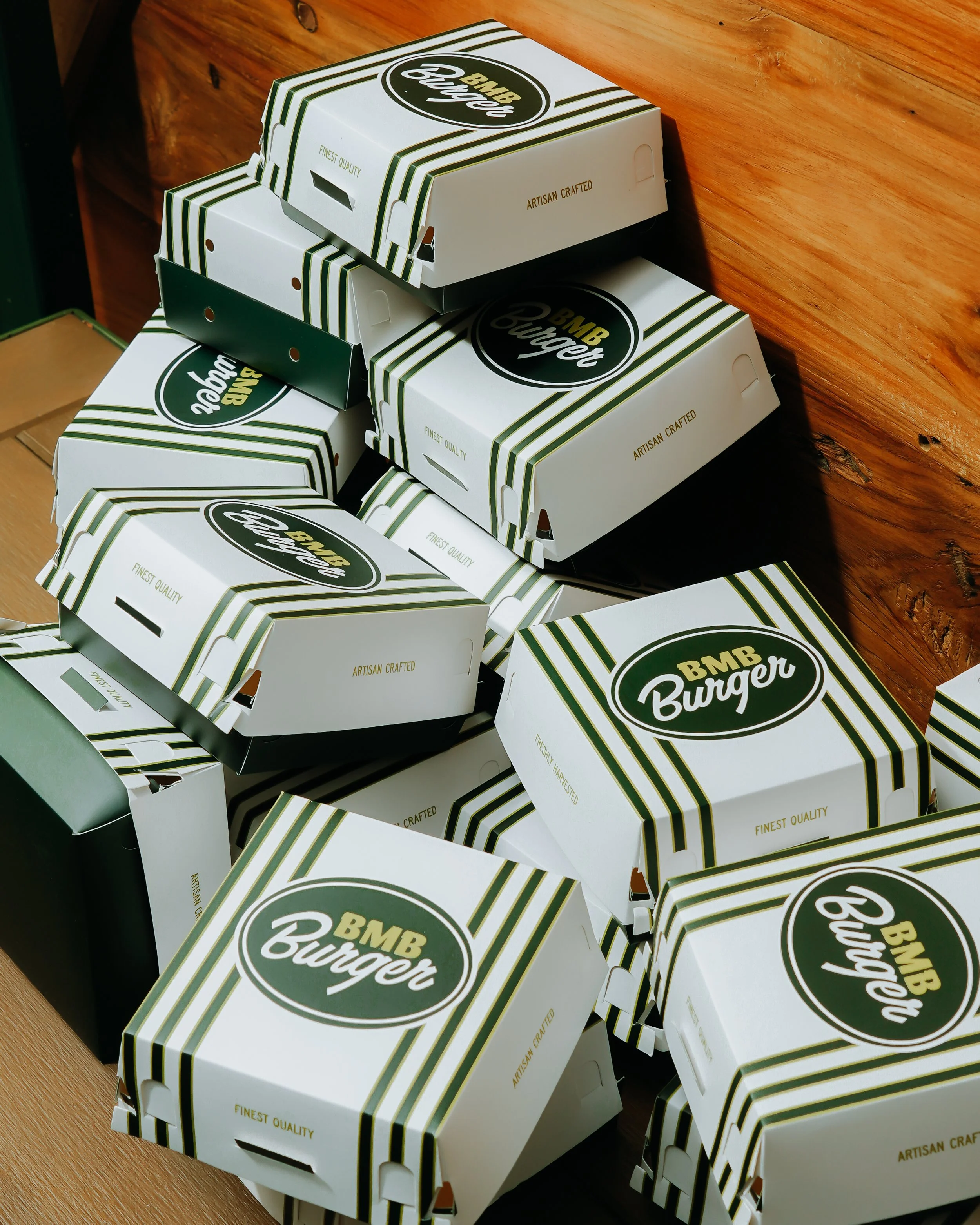 Multiple takeout burger boxes with green and white striped design and the logo 'BMB Burger' stacked on a wooden surface.