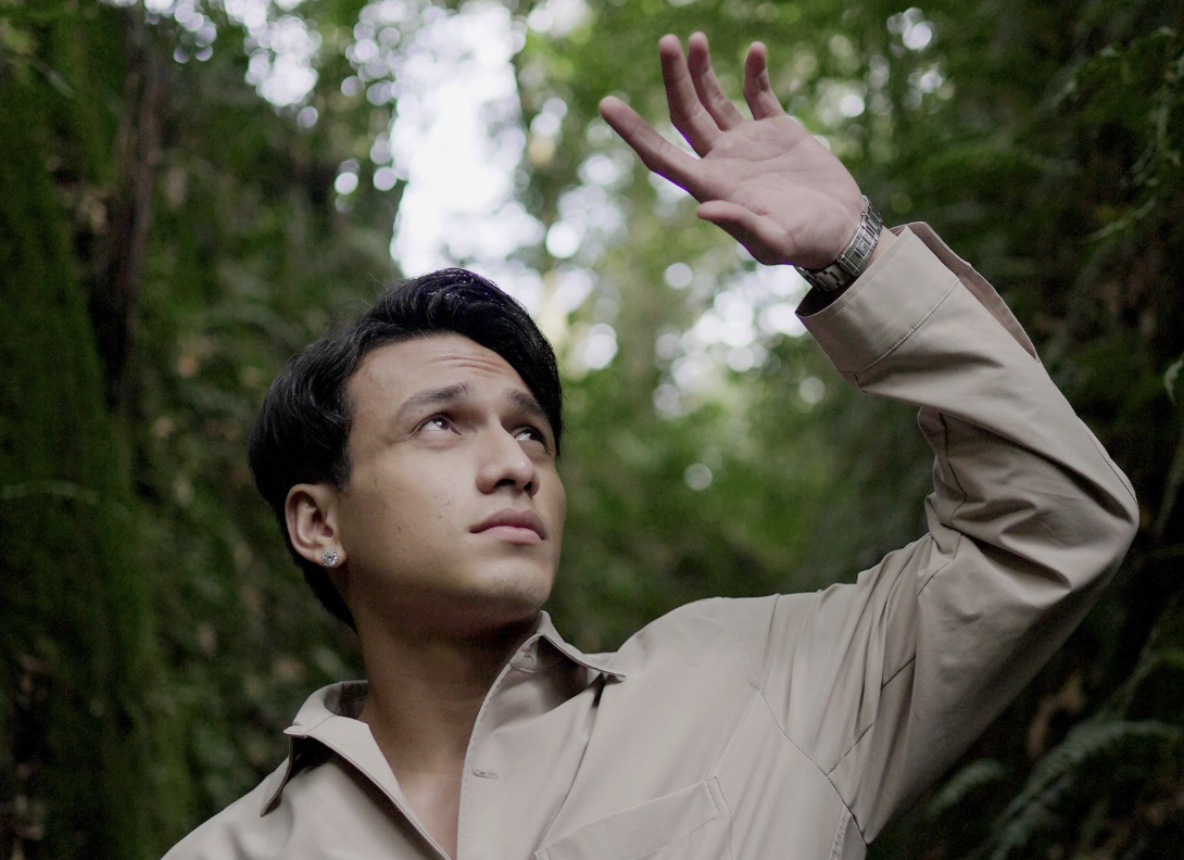 A man in a beige jacket raising his hand in a forest with green foliage.
