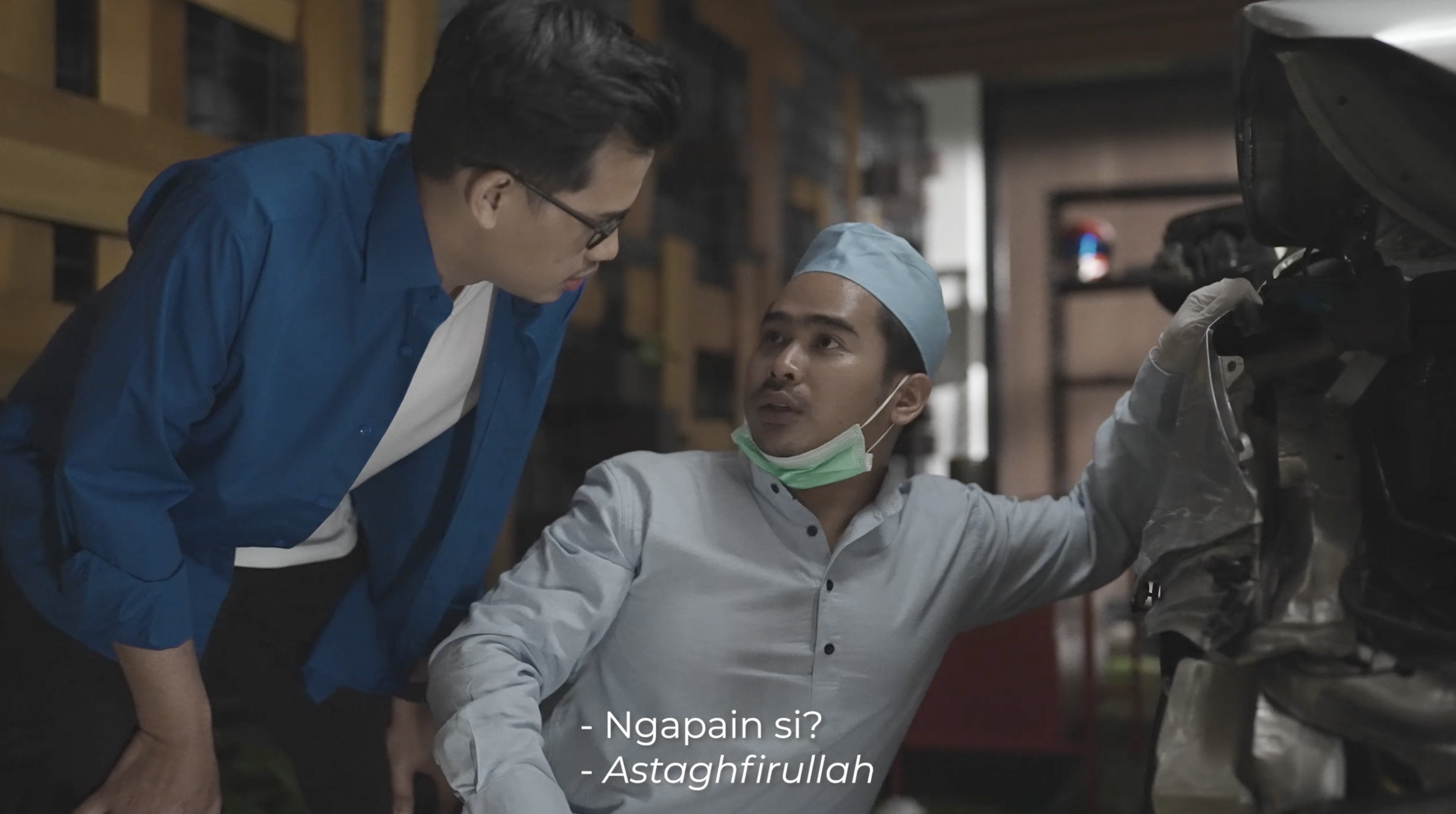 Two men, one wearing glasses and a blue shirt and another in a gray shirt and blue cap, are working on a car in a garage. The man in gray is holding a part of the car's engine, and the man in blue is leaning in to observe. There are subtitles that read, "Ngapain si?" and "Astaghfirullah."