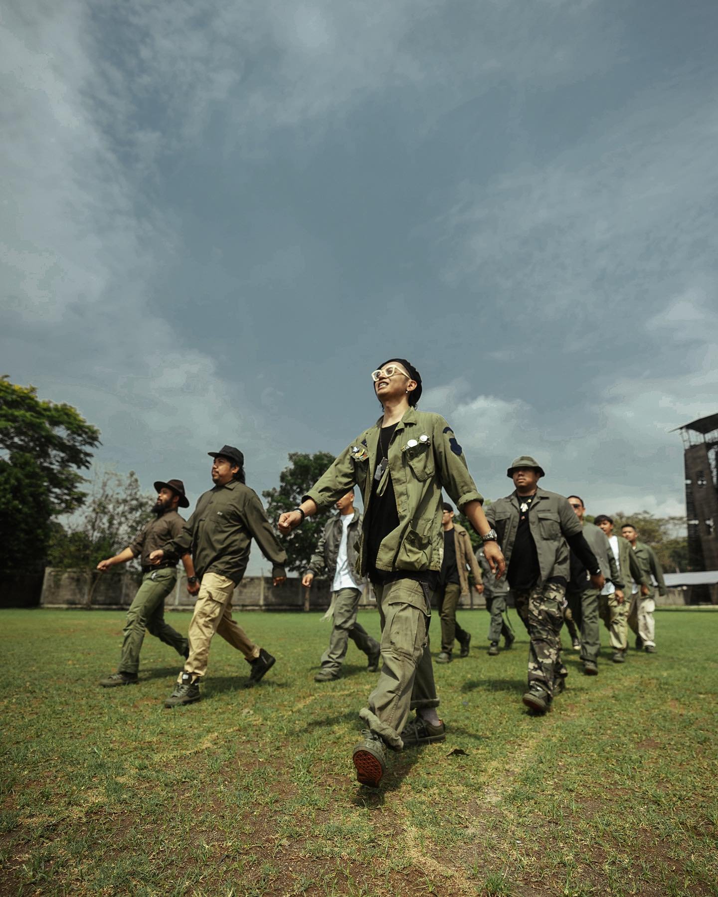 Group of people walking on a grassy field under cloudy sky, wearing casual and outdoor clothing.