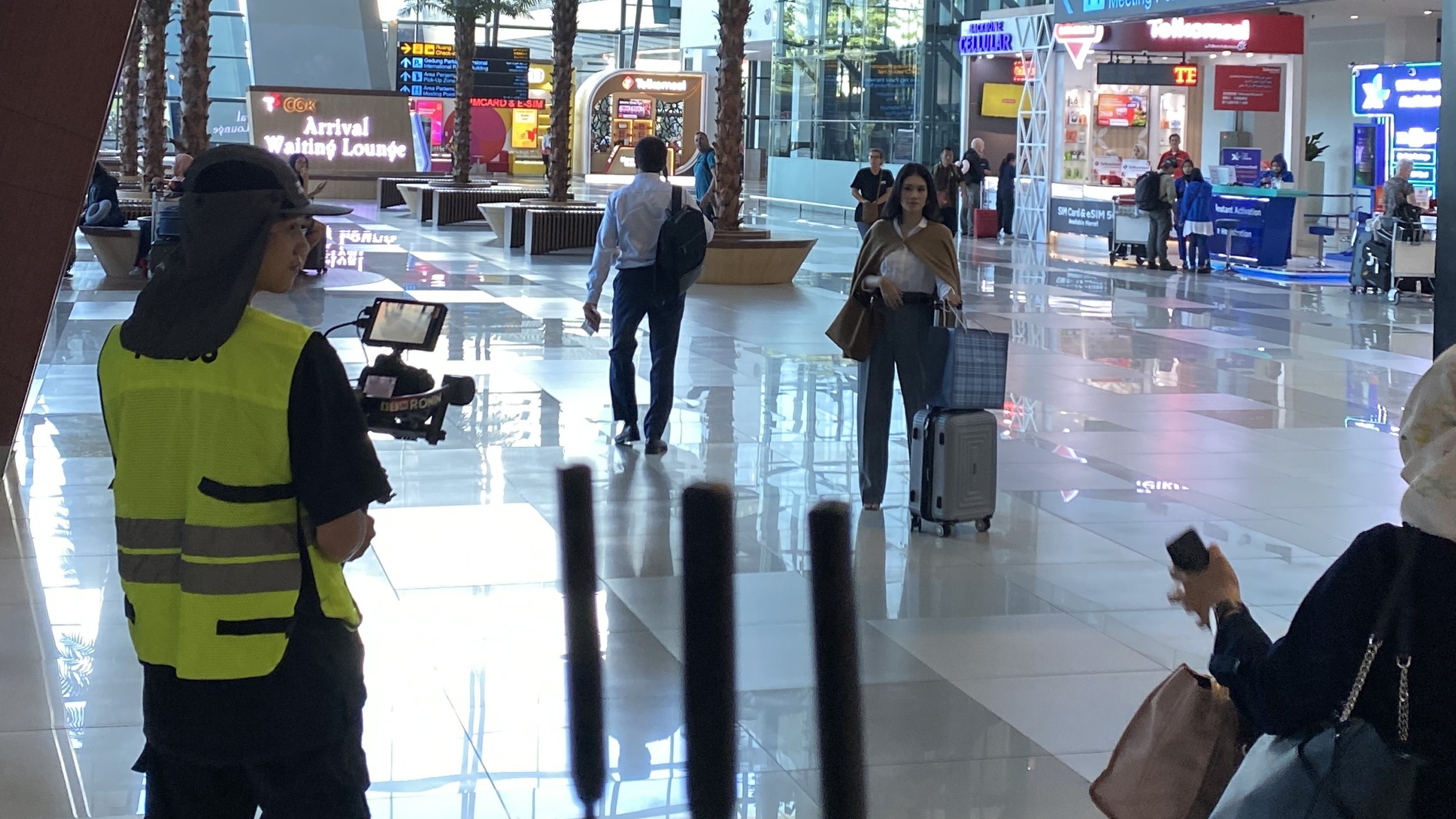 Passengers walking with luggage in an airport terminal, with digital signage and service counters in the background.