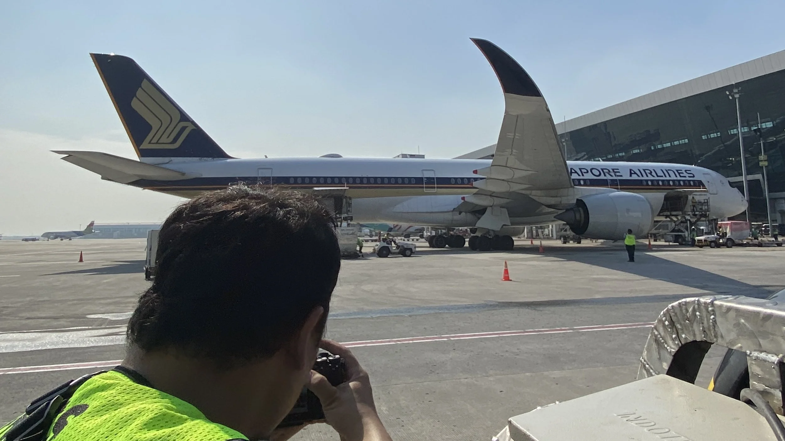 A Singapore Airlines airplane parked on the tarmac at an airport, with ground crew and a photographer in a fluorescent vest taking pictures in the foreground.