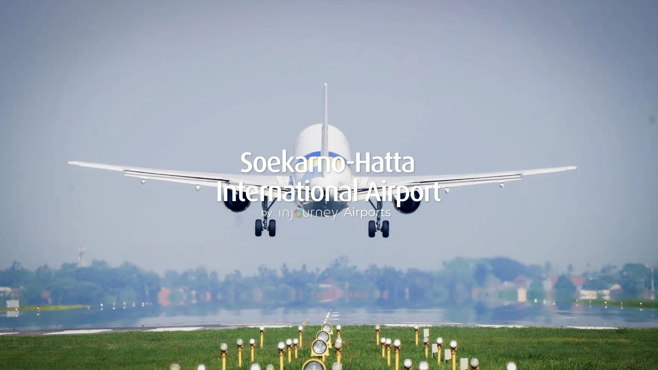 An airplane landing on a runway at Soekarno-Hatta International Airport, with lights lining the runway in the foreground and a cityscape in the background.
