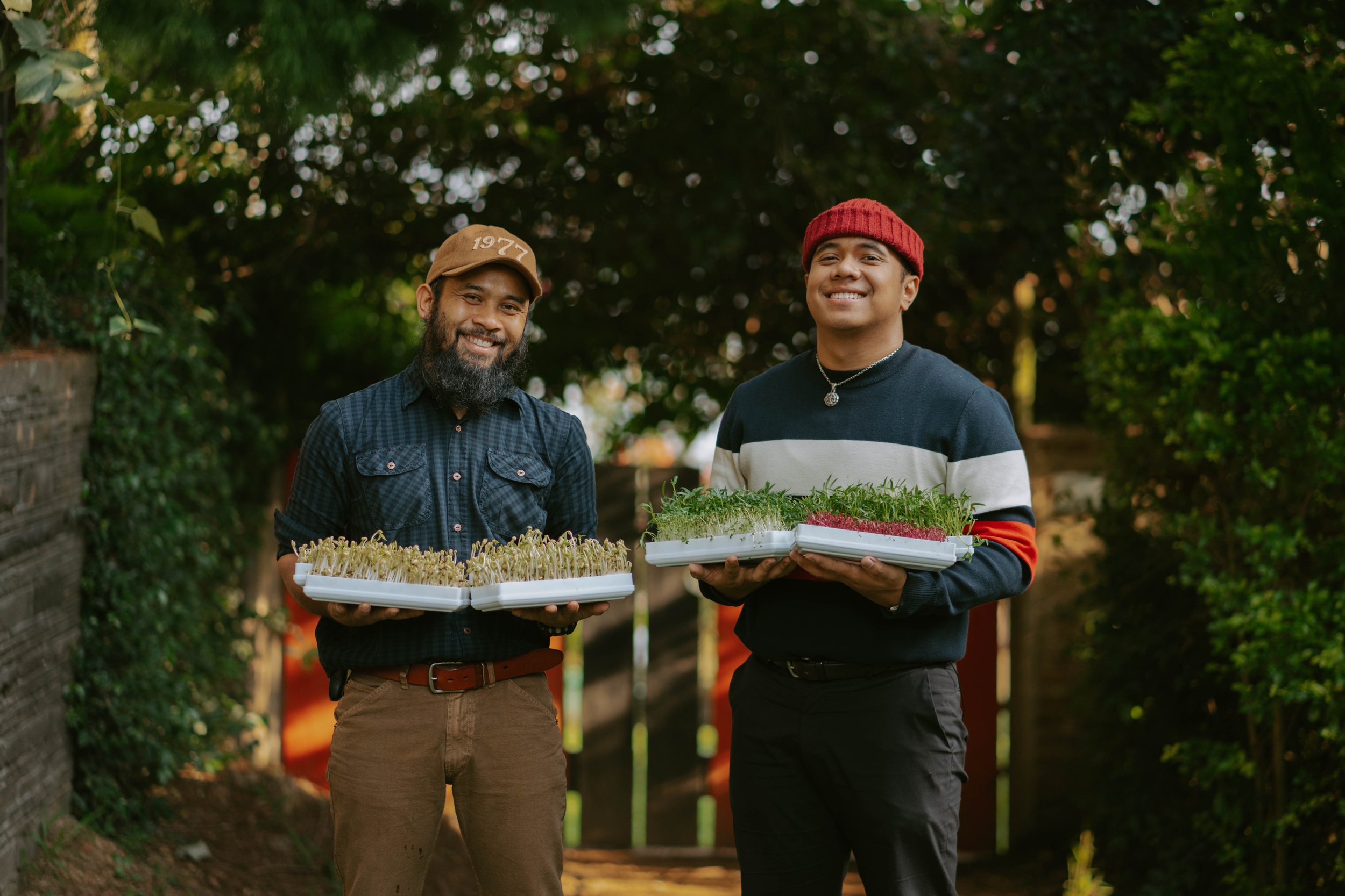 Two men smiling outdoors, holding trays of microgreens, with greenery and trees in the background.