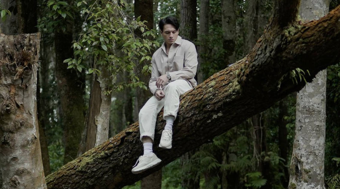 A young man sitting on a fallen tree in a forest, surrounded by green trees and foliage.