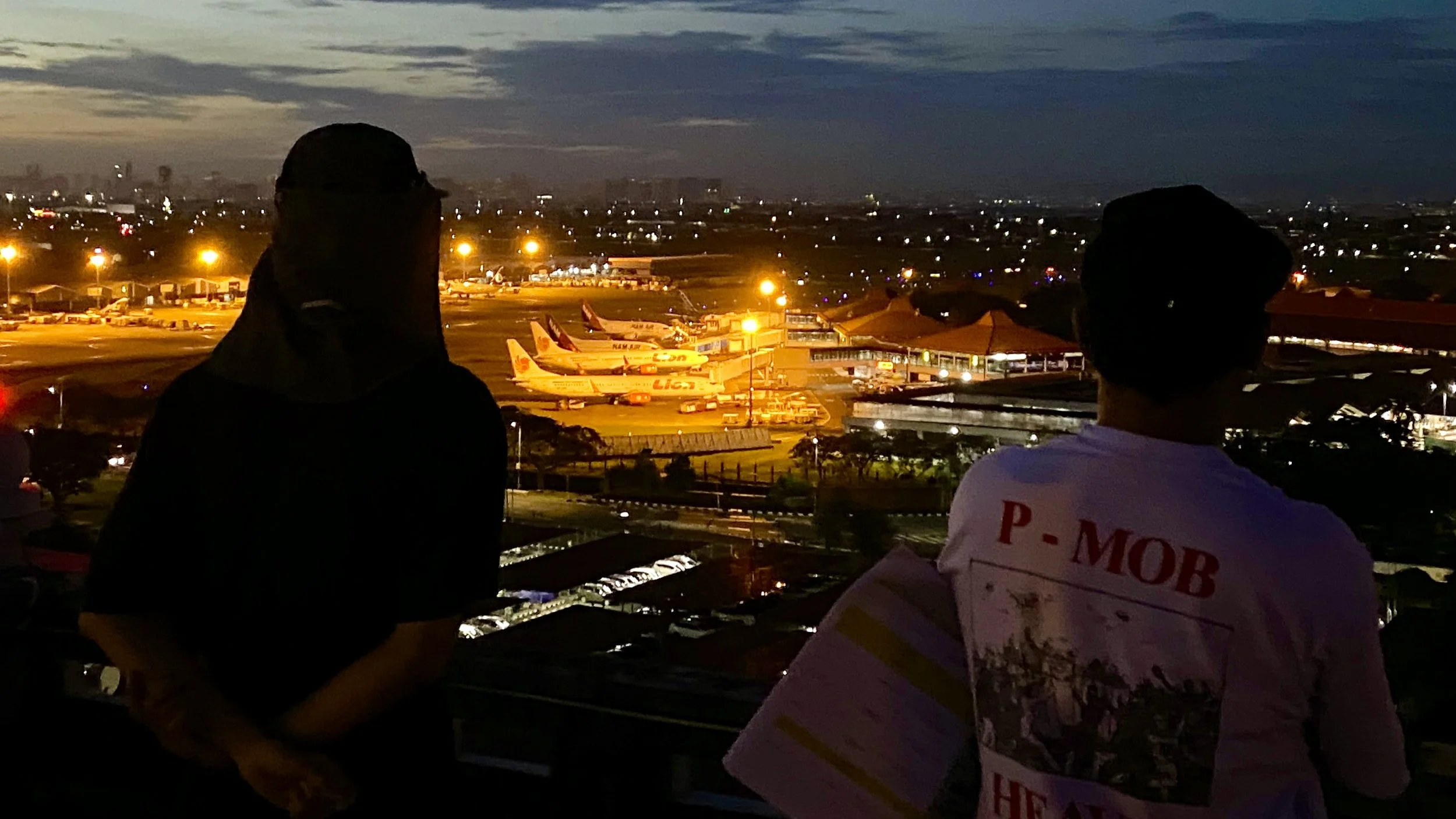 Two silhouetted individuals standing and looking over an airport tarmac at dusk, with several airplanes parked and illuminated buildings in the background.