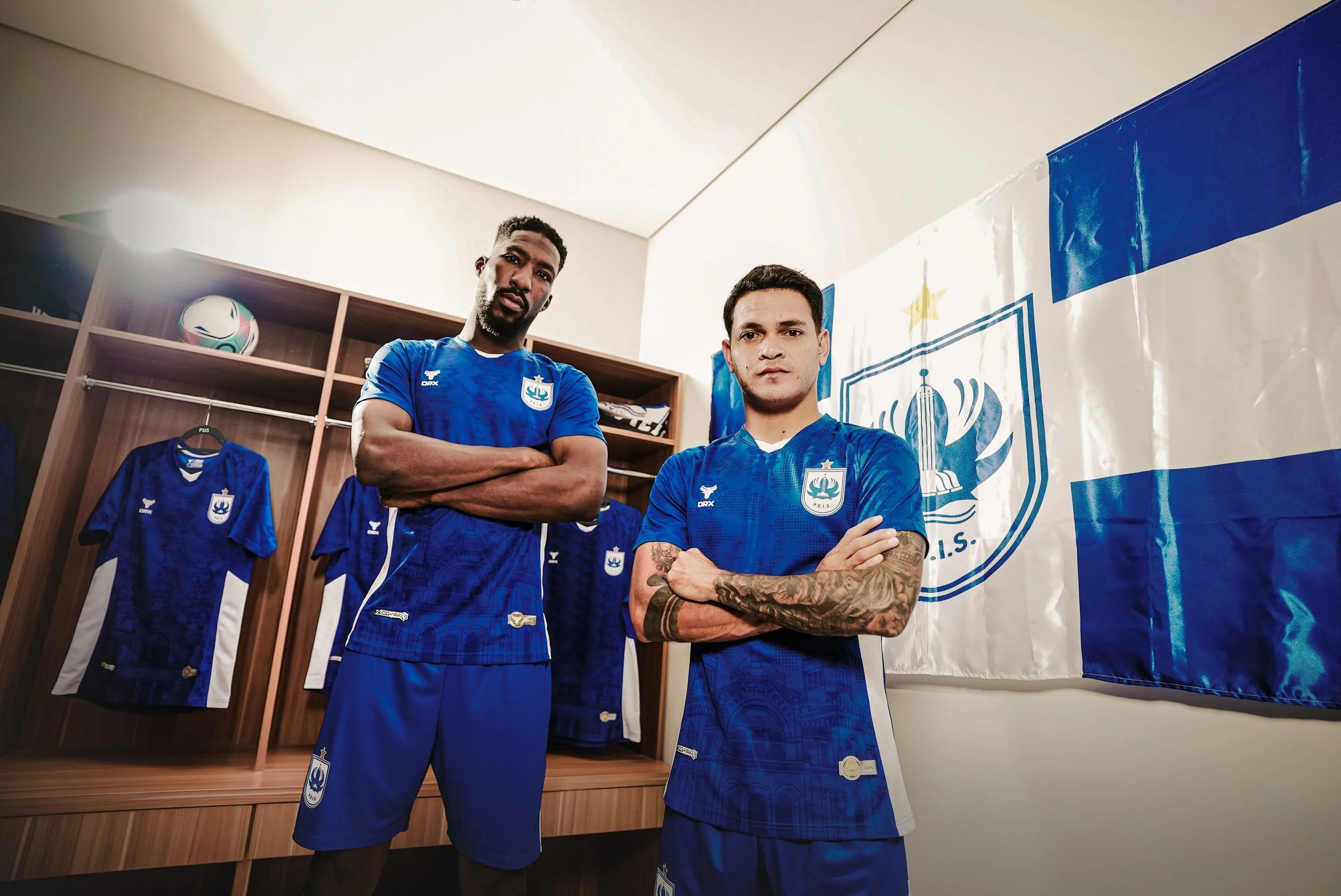 Two male soccer players standing with arms crossed inside a locker room, with blue jerseys and a large banner with a crest on the wall behind them.