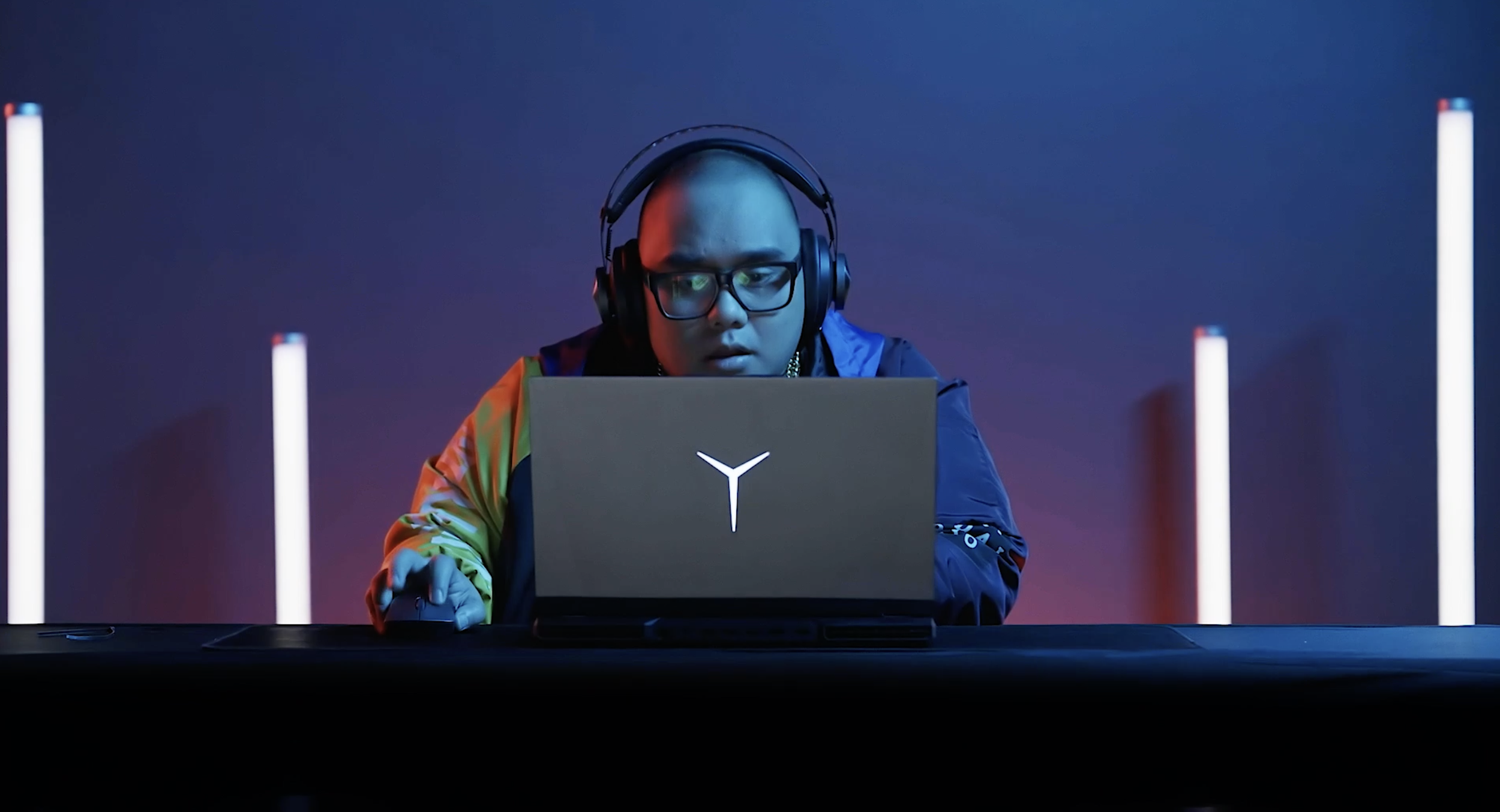 A person wearing glasses and headphones, sitting at a desk with a laptop that has a white logo on the back, under colorful lighting with vertical white light bars in the background.
