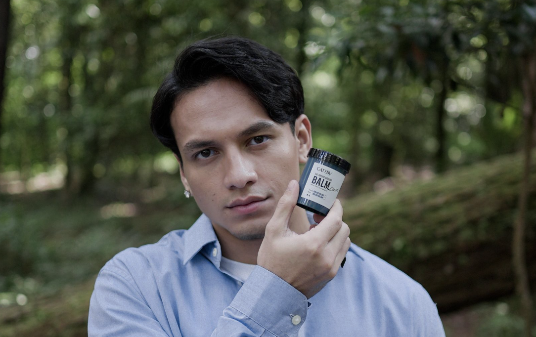 Young man outdoors holding a small black jar labeled 'BALM'