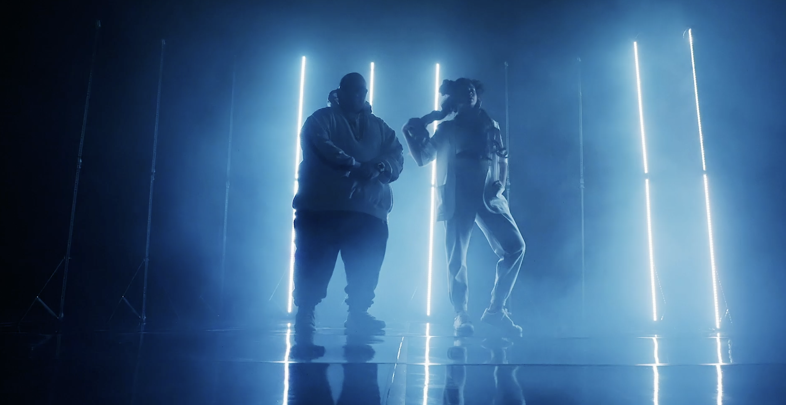 Two people standing on a stage with backlighting, silhouetted against vertical blue light strips, creating a futuristic or artistic atmosphere.