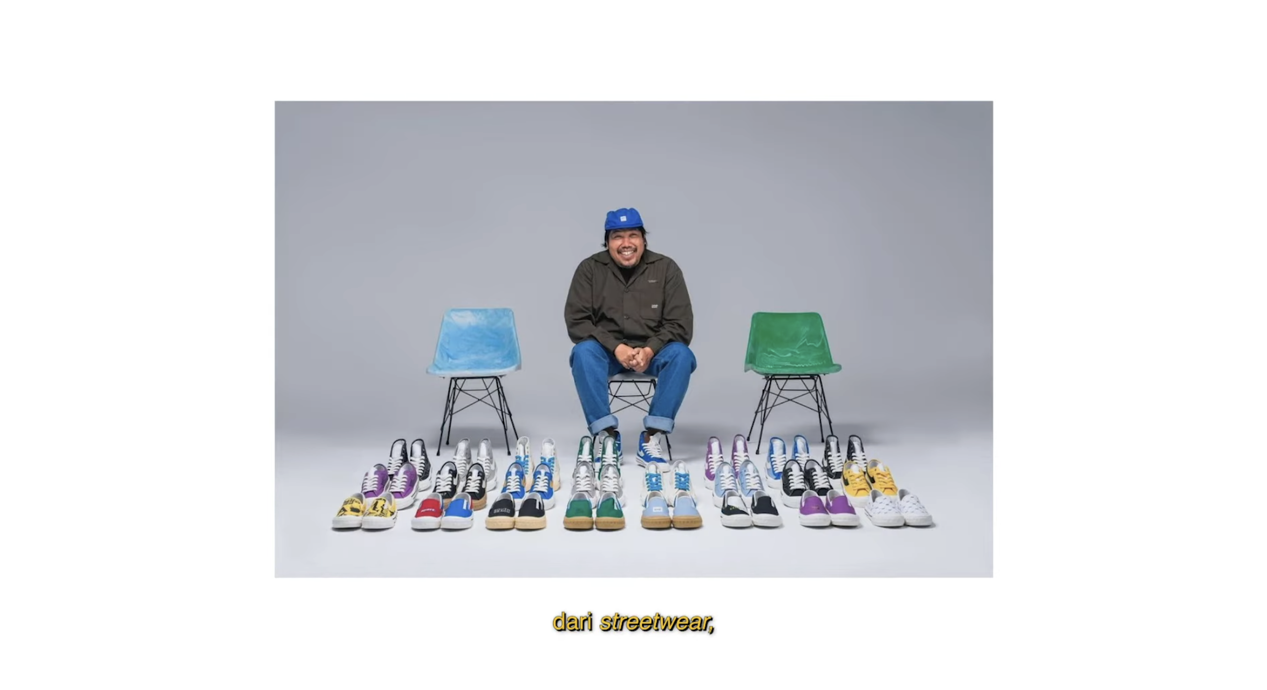 A man with a blue hat sitting on a black chair surrounded by colorful sneakers arranged in pairs on the floor, with two empty chairs, one blue and one green, on either side of him, set against a gray background.