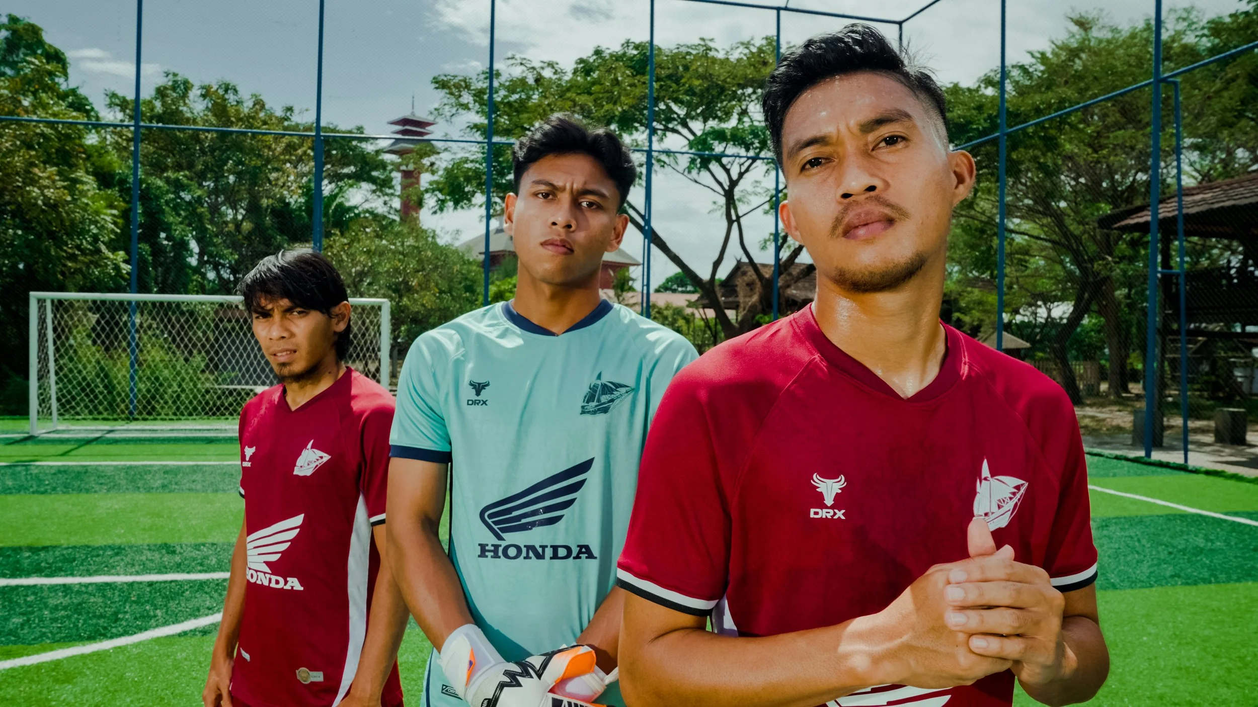 Three young men standing on a soccer field during daytime, looking at the camera with serious expressions. Two wear red soccer jerseys, one wears a light blue goalkeeper jersey with a Honda logo, in front of a goalpost and green artificial turf, with trees and blue sky in the background.