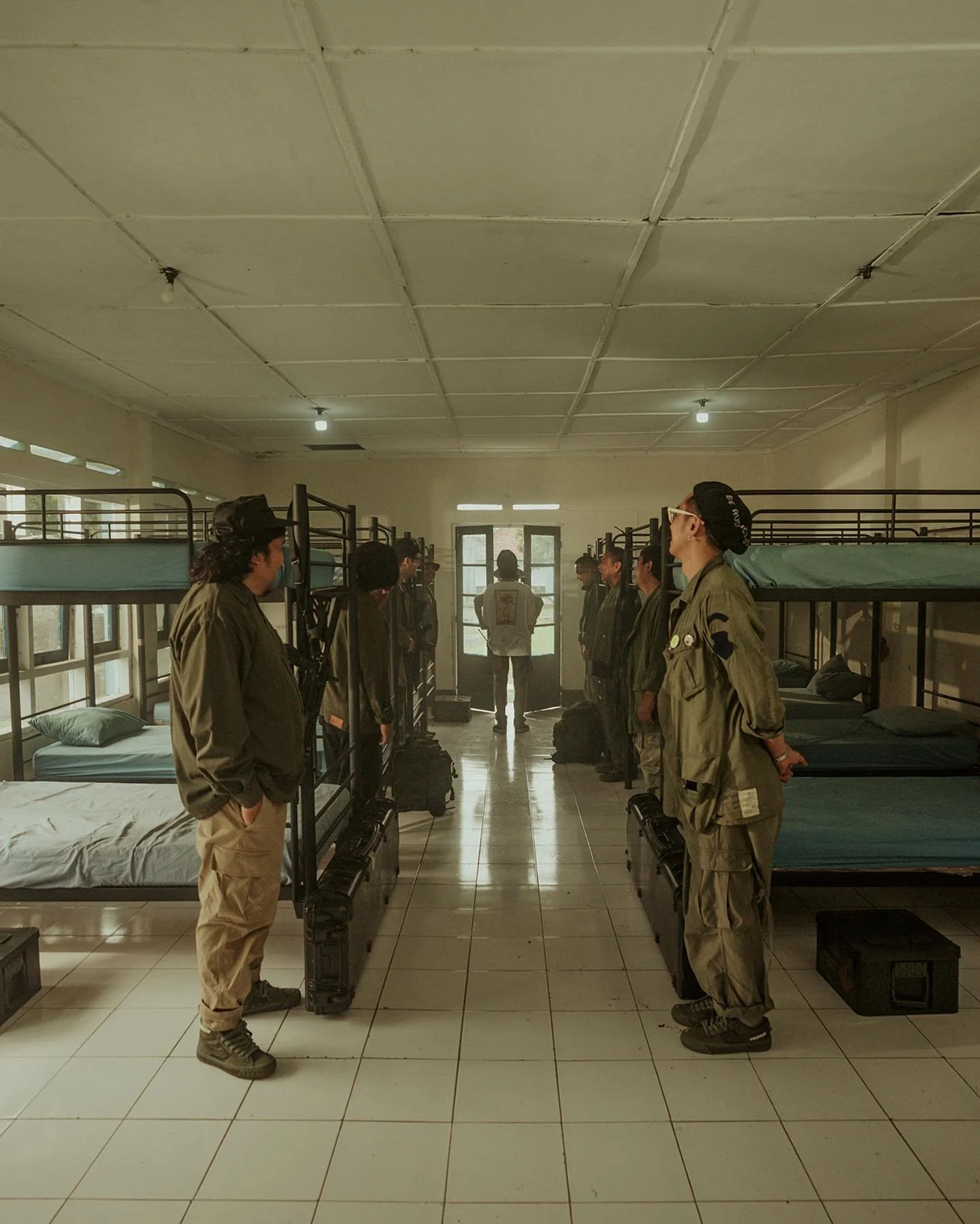 A group of people standing in uniform on each side of a corridor in a dorm room with bunk beds, with a person sitting on a chair at the far end near the door.