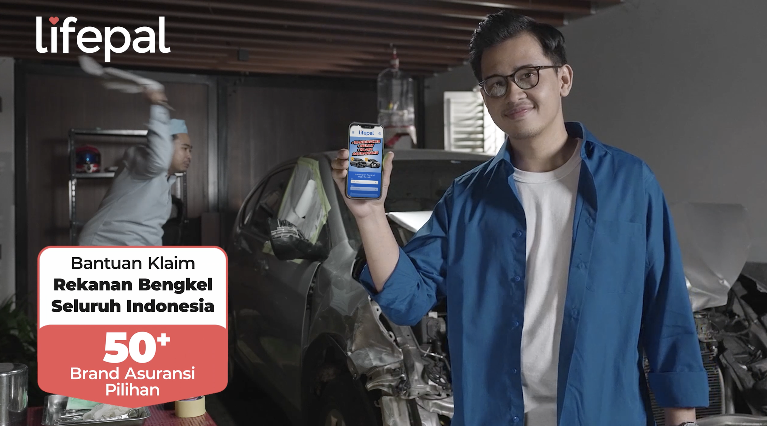 A young man in glasses and a blue shirt holds up a smartphone displaying the Lifepal app, standing in front of a damaged car in a garage with a mechanic working in the background. There is a promotional banner in Indonesian mentioning insurance claims and options.