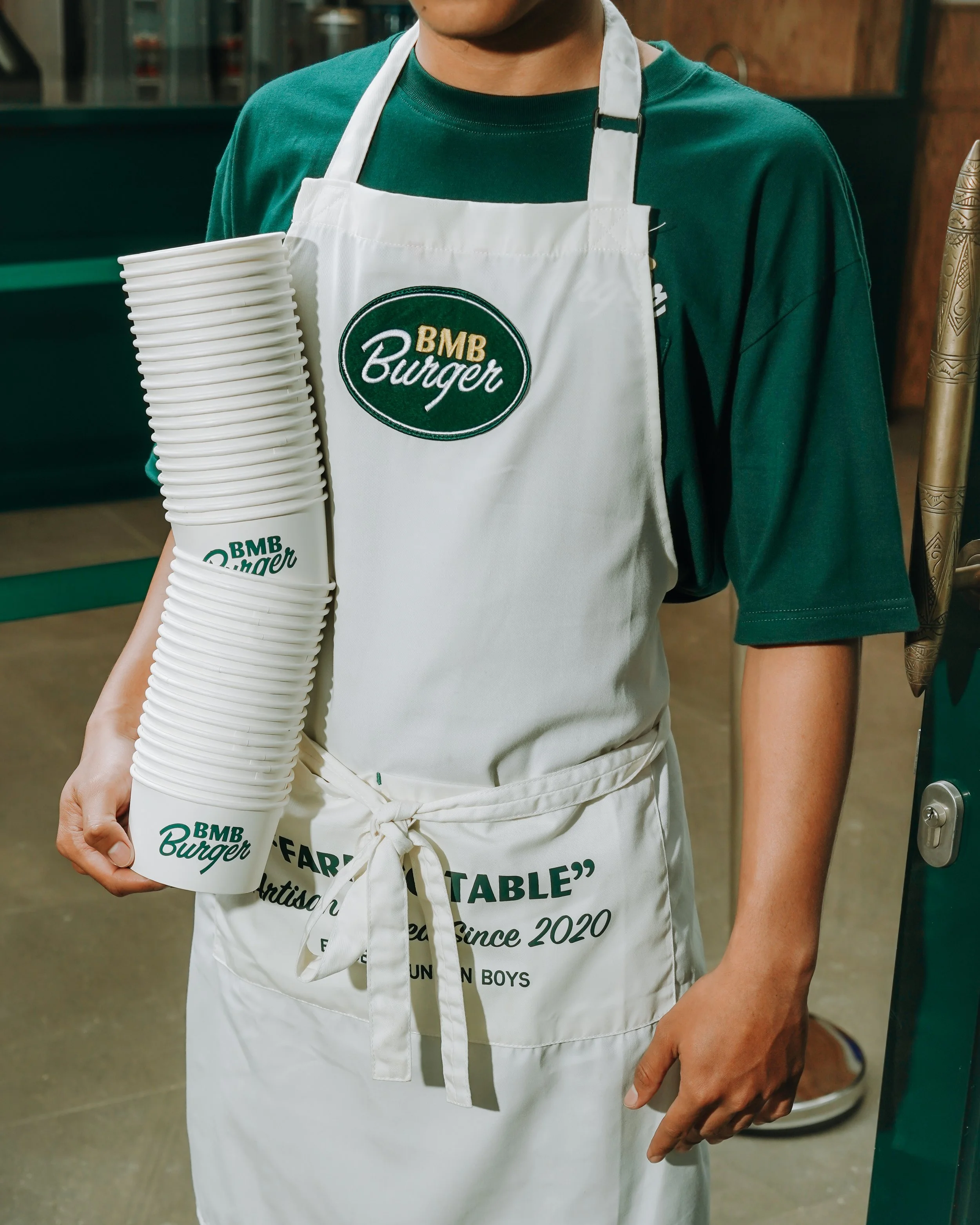 Person wearing a green shirt and white apron holding a stack of paper plates with 'BMB Burger' logo.