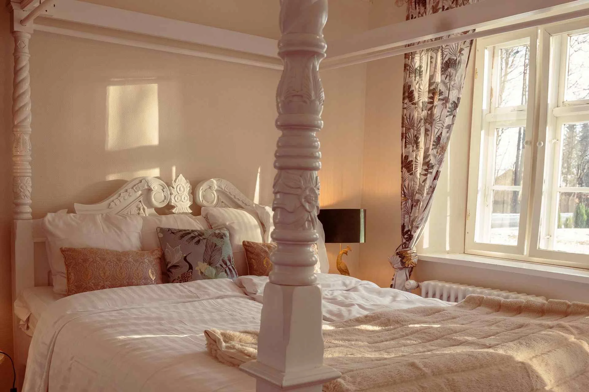 Et soveværelse med en stor seng, dekorative puder og udskåret sengegavl. Store vinduer med blomster gardiner og et sidebord med en sort lampe.