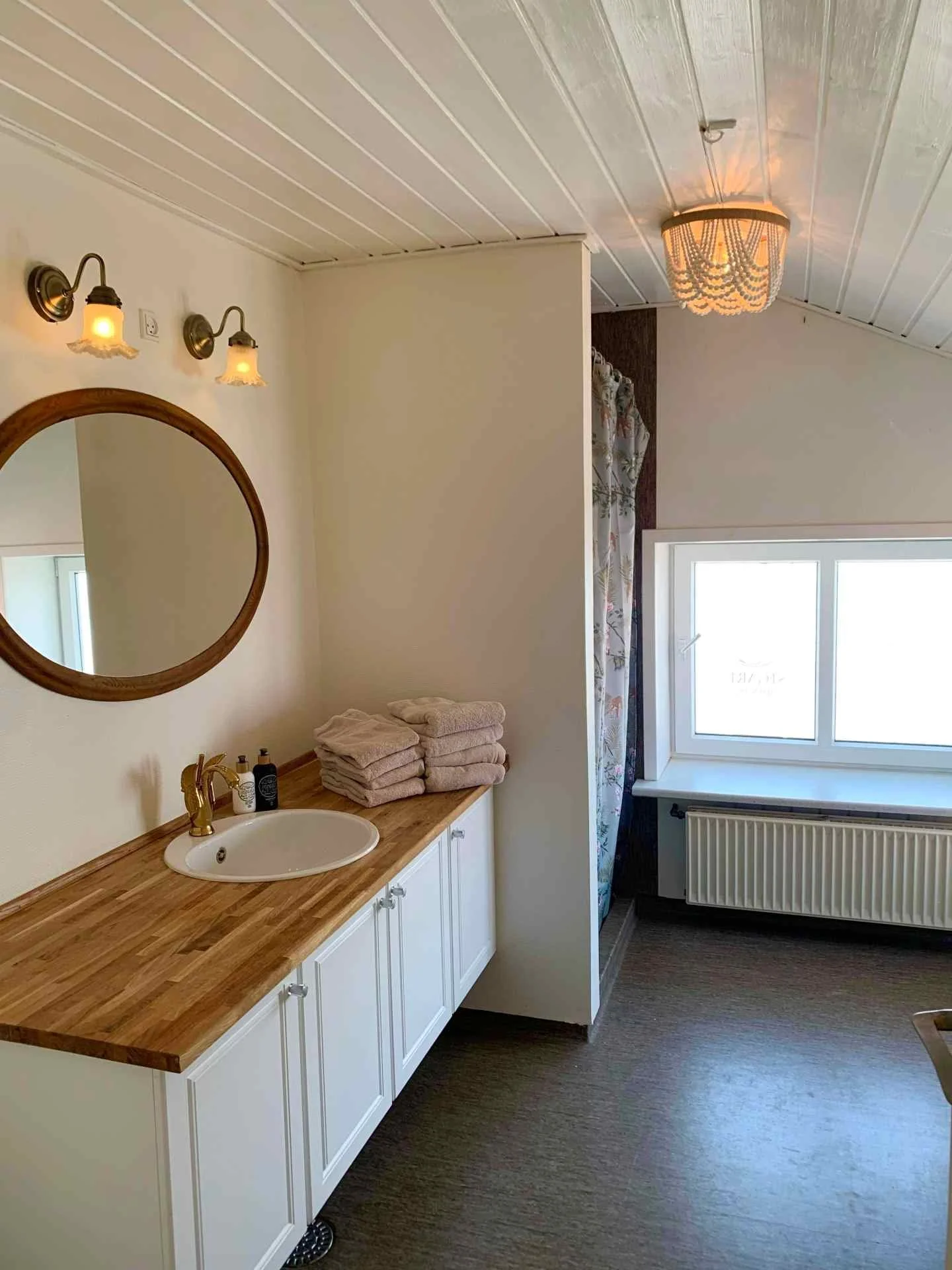 Badeværelse med hvidt skab med træbordplade, håndklæder, round spejl, væglampe, vindue og radiator.