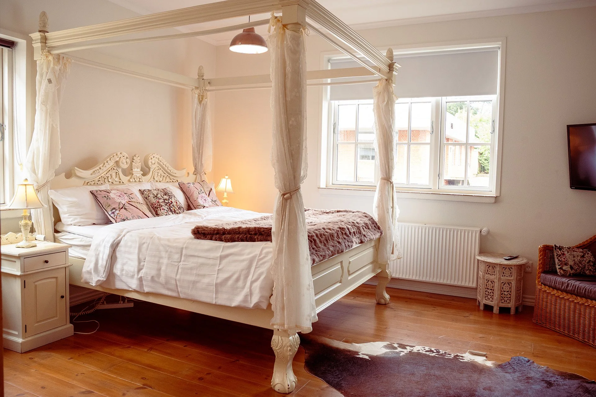 Et soveværelse med en hvid fire-søjlet seng, pyntede med puder og et tykt, blushfarvet fleece tæppe. Der er to natbord modeller med små lamper, naturlige trægulve, en TV på væggen, og store vinduer, der lader lys ind i rummet.