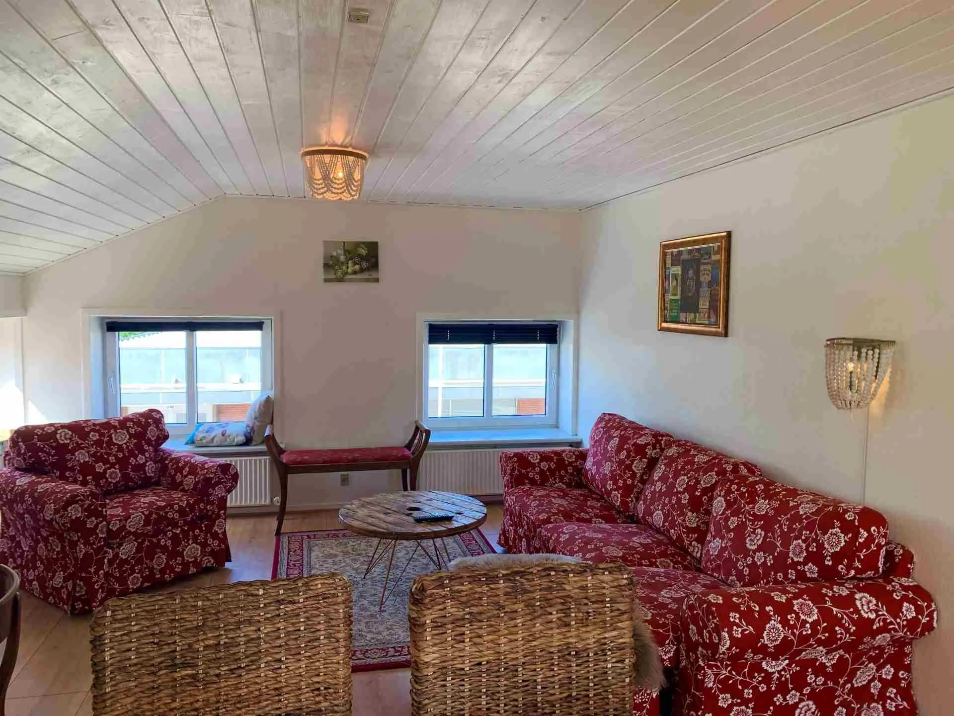 Stue med to store vinduer, to røde floral sofaer, en bænket, en rund træbord, et lamper og nogle vægdekorationer.